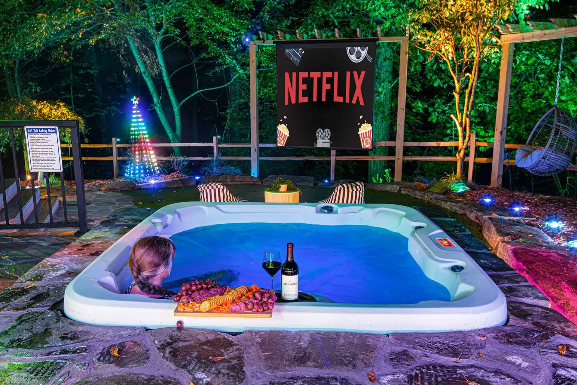 Woman relaxes in a hot tub with wine and snacks next to a Netflix screen in a backyard.