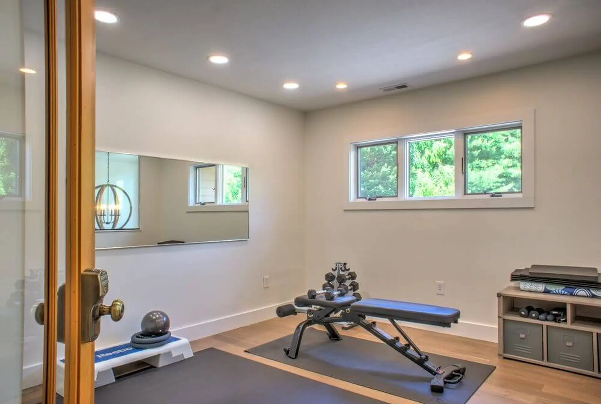 Gym equipment, including a weight bench and dumbbells, is shown in a room with a mirror and windows.