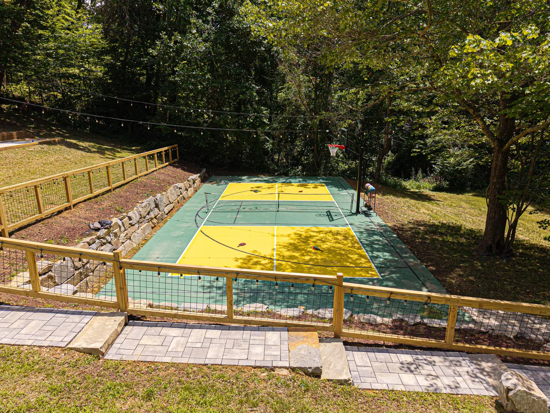 An outdoor basketball court with a tennis net, surrounded by trees and a wooden fence with lights.