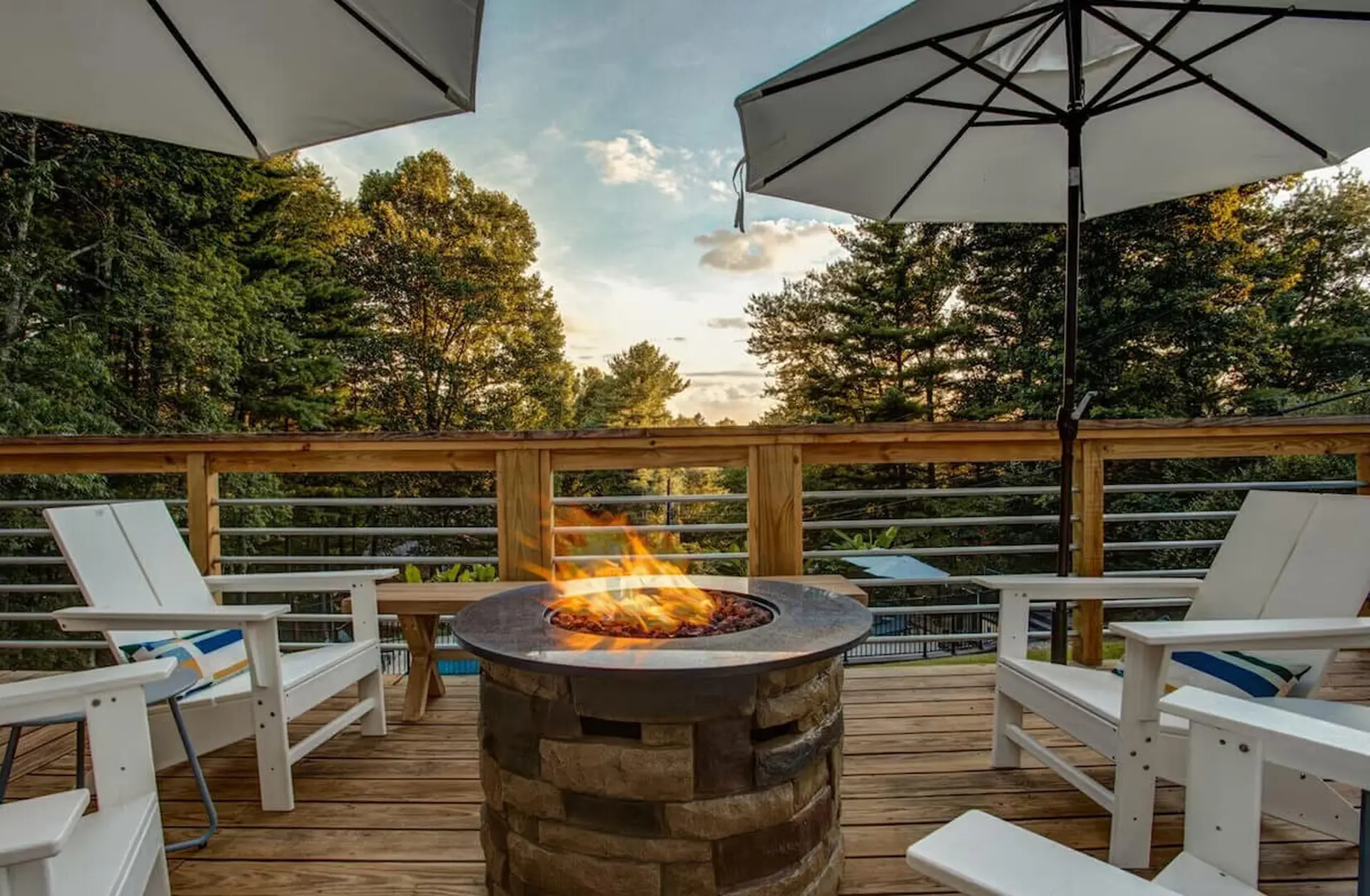 A deck with a fire pit, chairs, and umbrellas overlooks a wooded area.