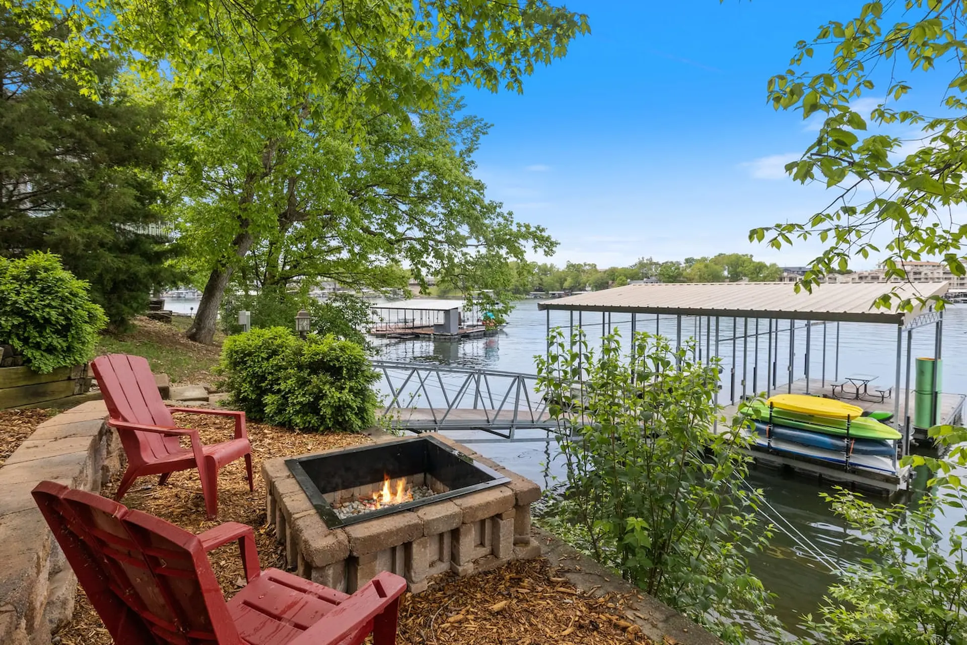 A fire pit with two chairs is on a patio overlooking a lake with a boathouse and kayaks.