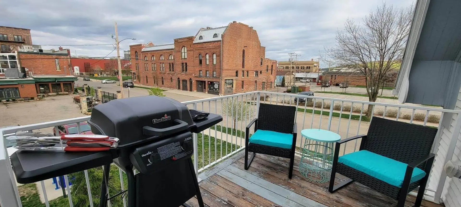 Balcony with a grill, chairs, and table overlooks a street with brick buildings.