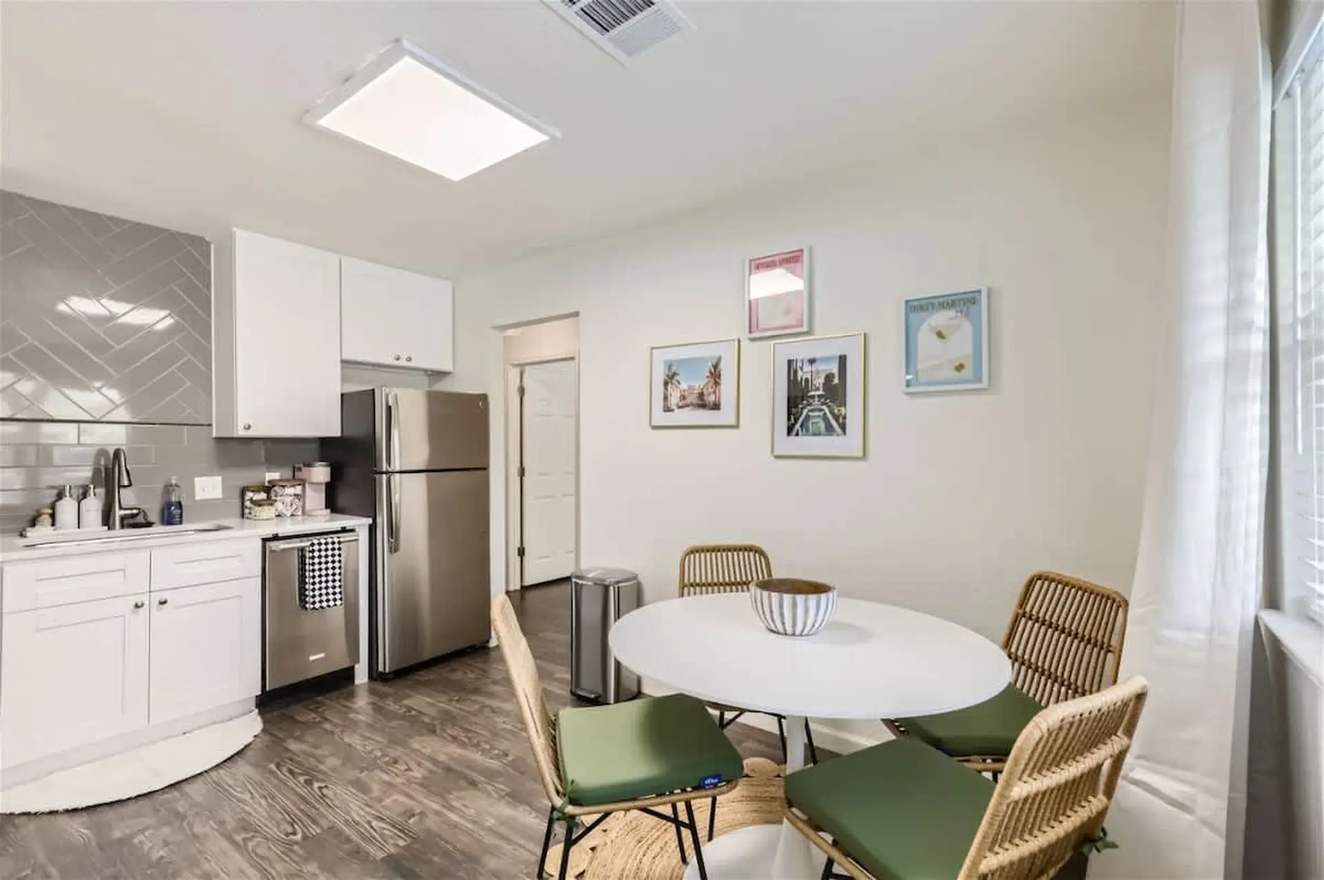 A dining area next to a kitchen with a white table, rattan chairs, and a steel refrigerator.