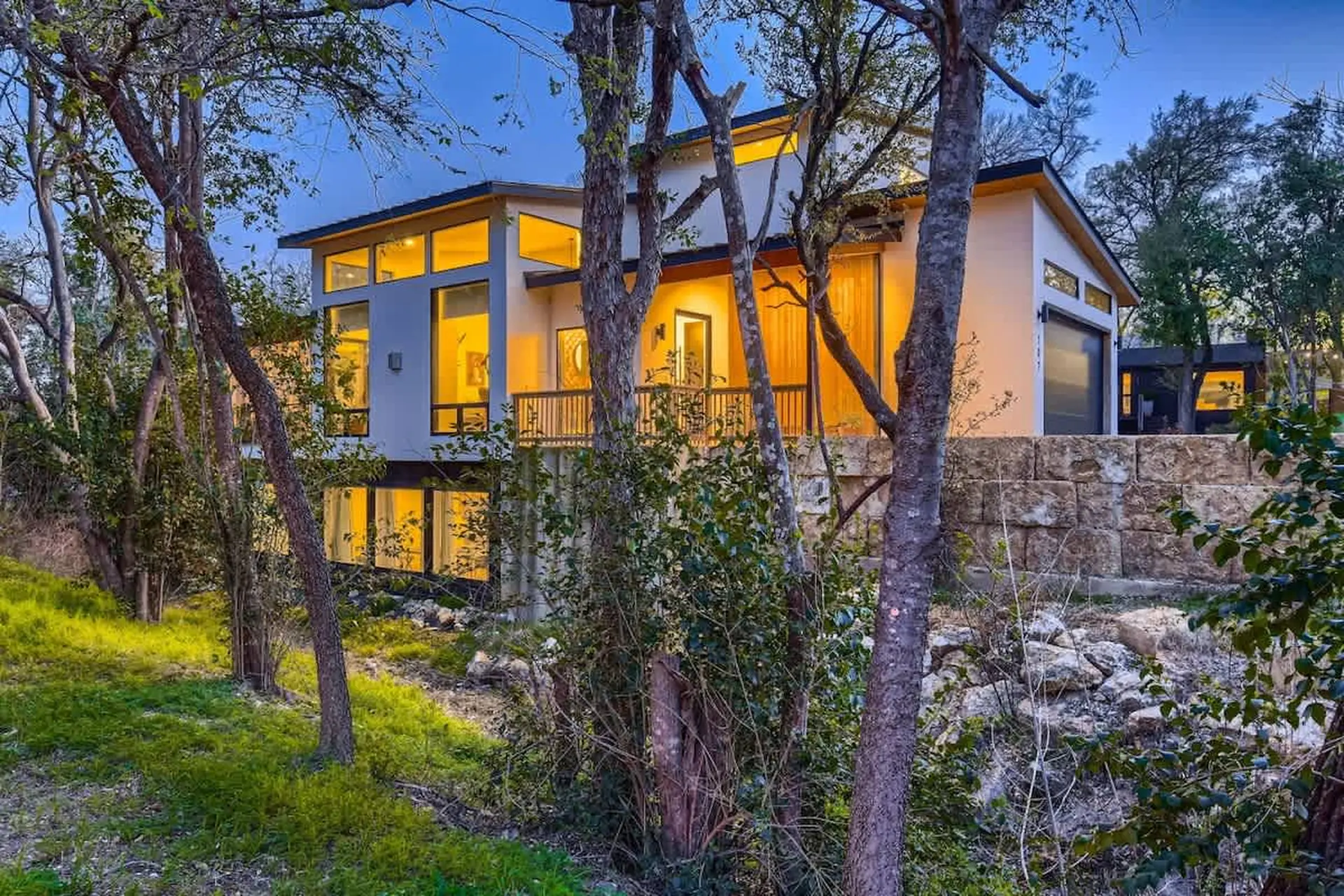 Modern home with large windows surrounded by trees and a stone wall at twilight.