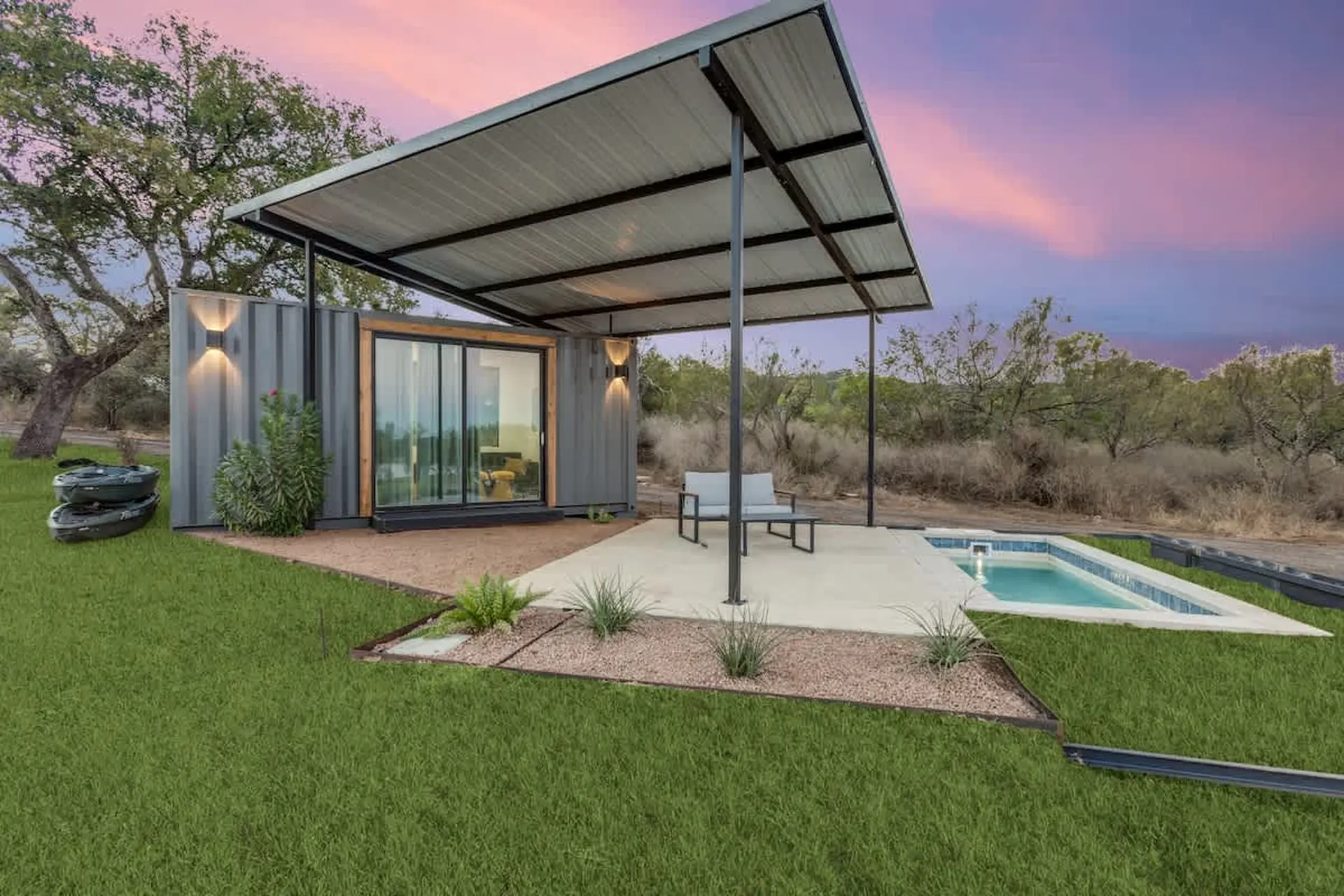 Modern shipping container home with a patio, small pool, and kayaks at sunset.
