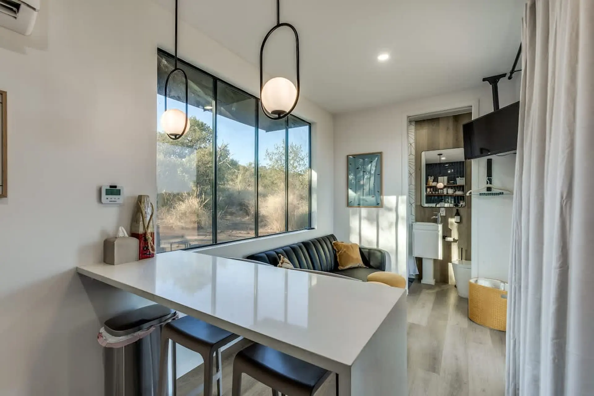 A modern living area with a white counter, black couch, and large window overlooking trees.