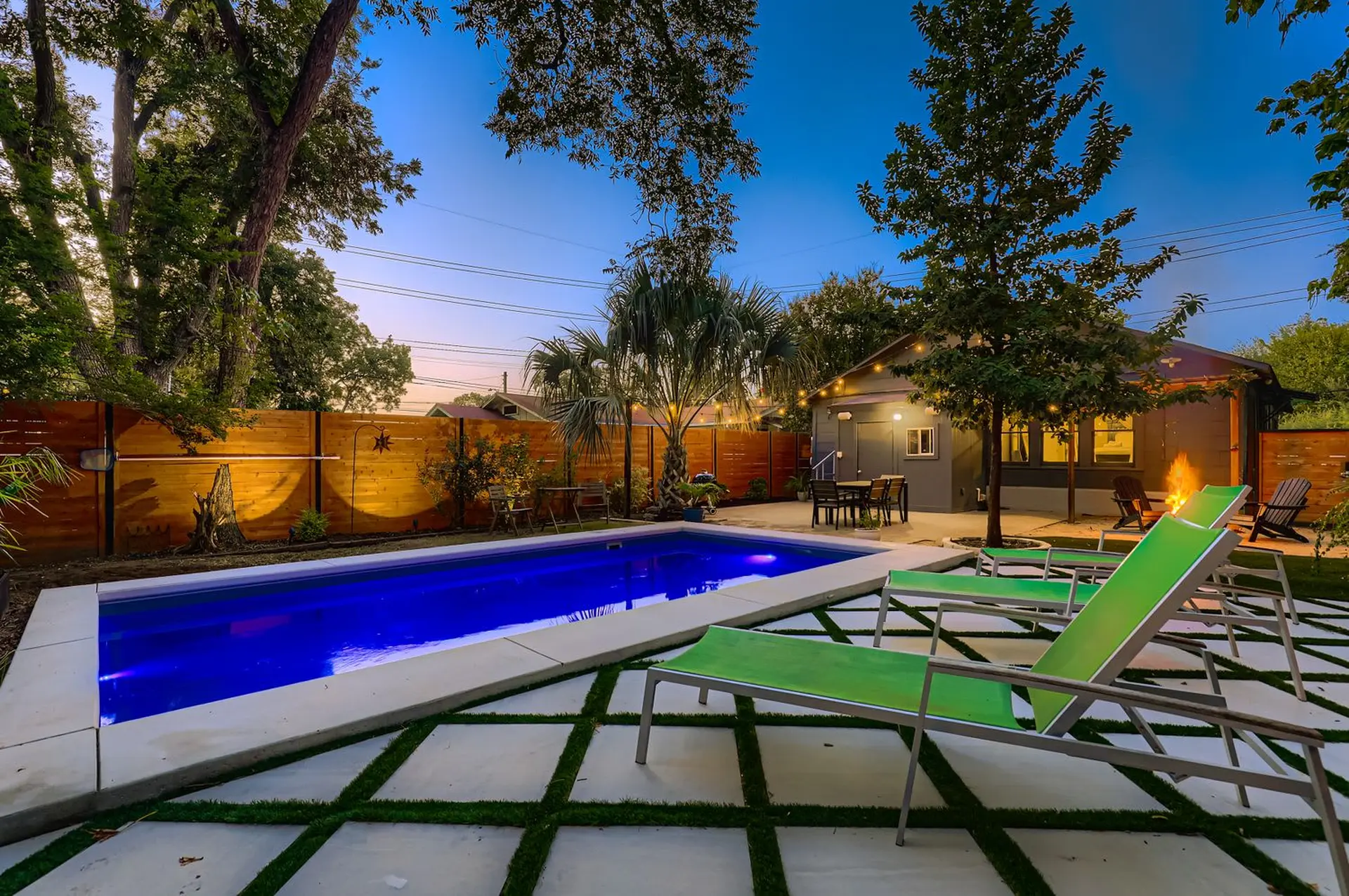 A backyard with a bright blue pool, lounge chairs, and a fire pit at dusk.