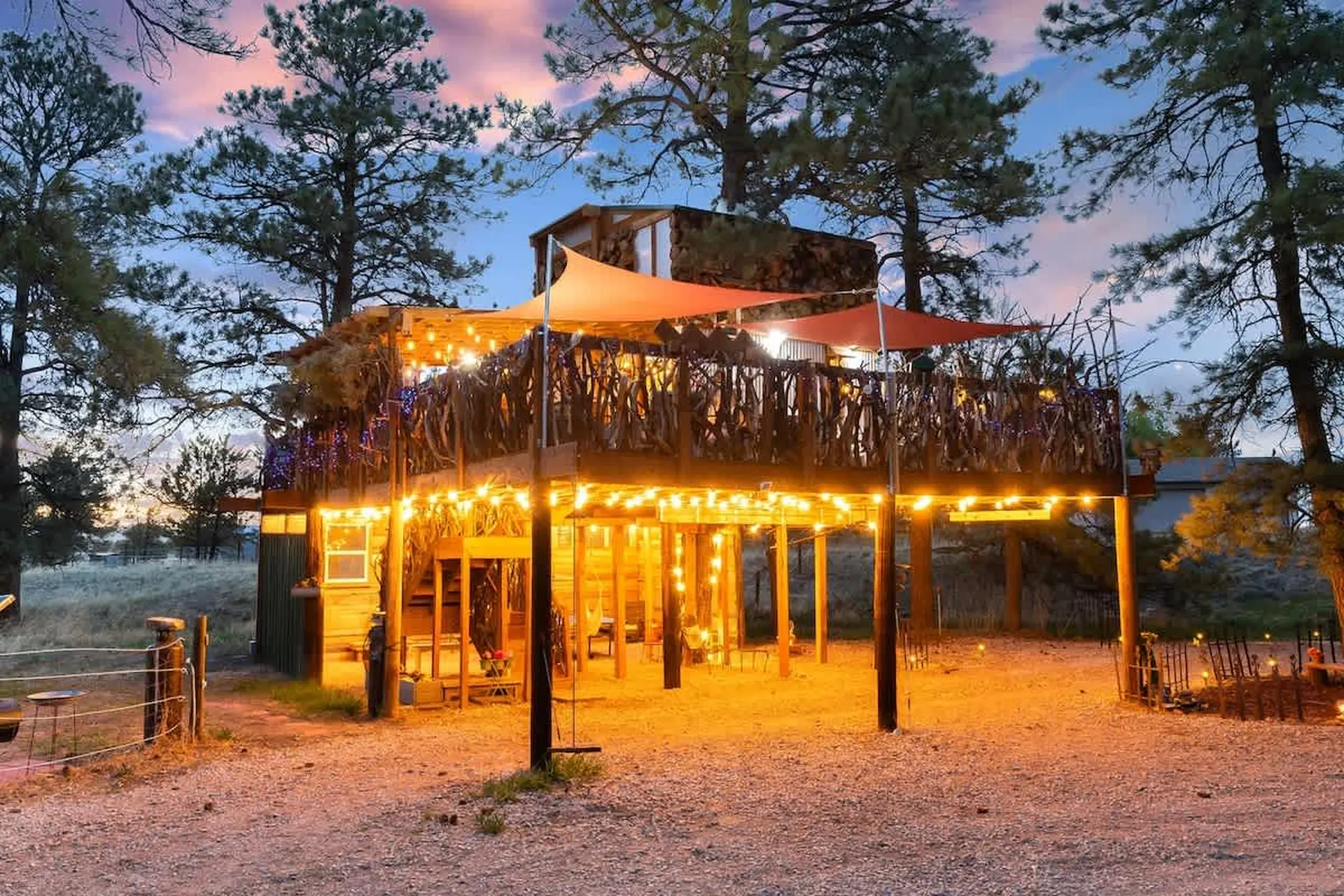 A rustic treehouse decorated with fairy lights and orange sunshades, nestled amongst pine trees at d