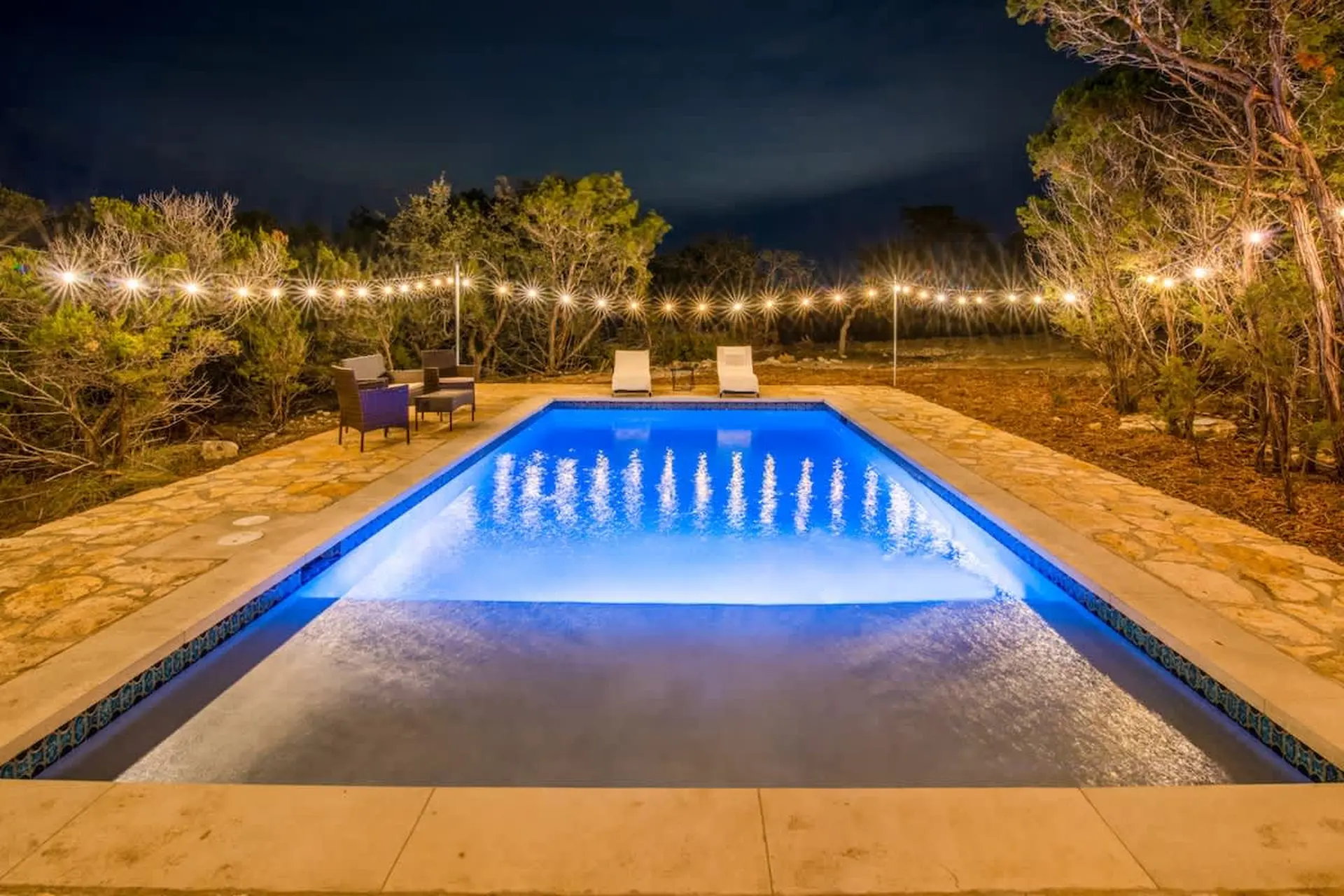 Outdoor pool at night with string lights and lounge chairs.