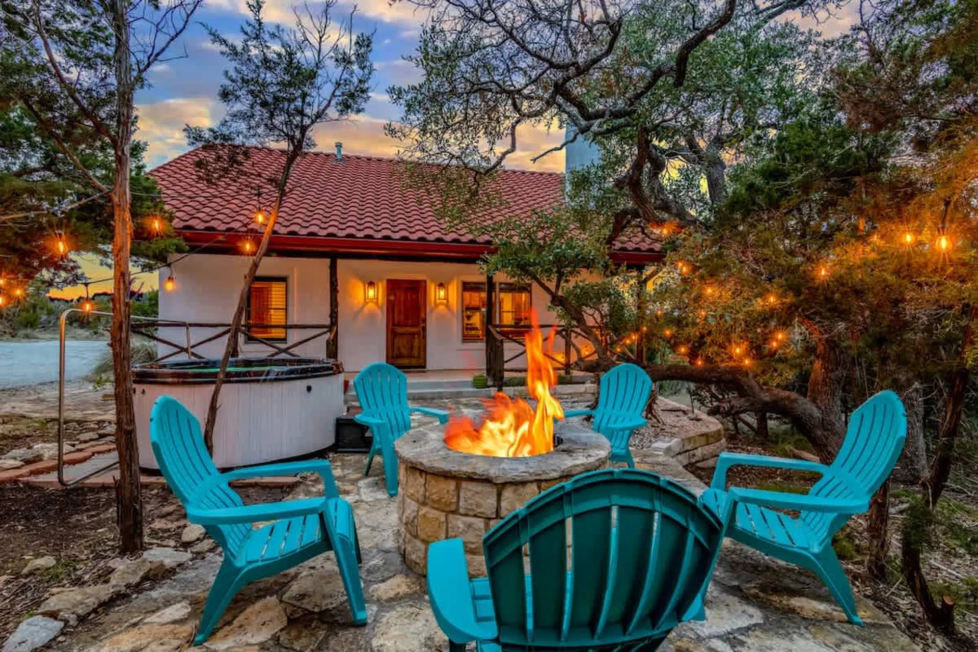 A cozy outdoor seating area with a fire pit, hot tub, and turquoise chairs under string lights.