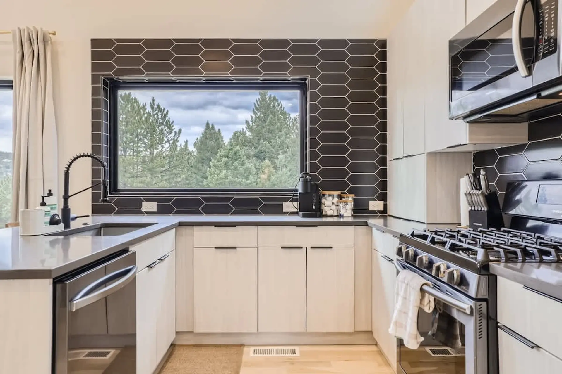A modern kitchen with a black hexagon tile backsplash and a large window overlooking trees.