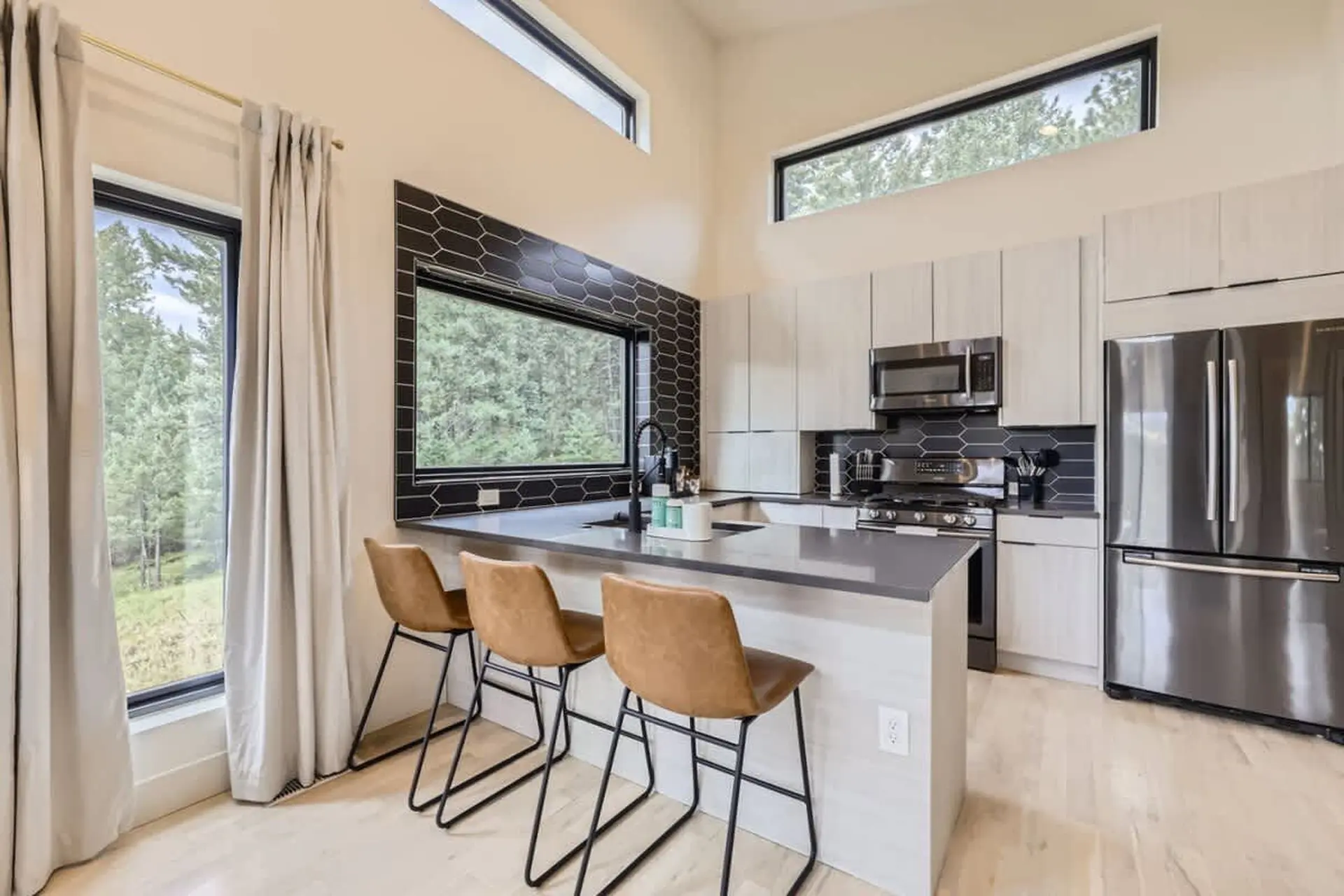 Modern kitchen with island, bar stools, and stainless steel appliances, featuring large windows.