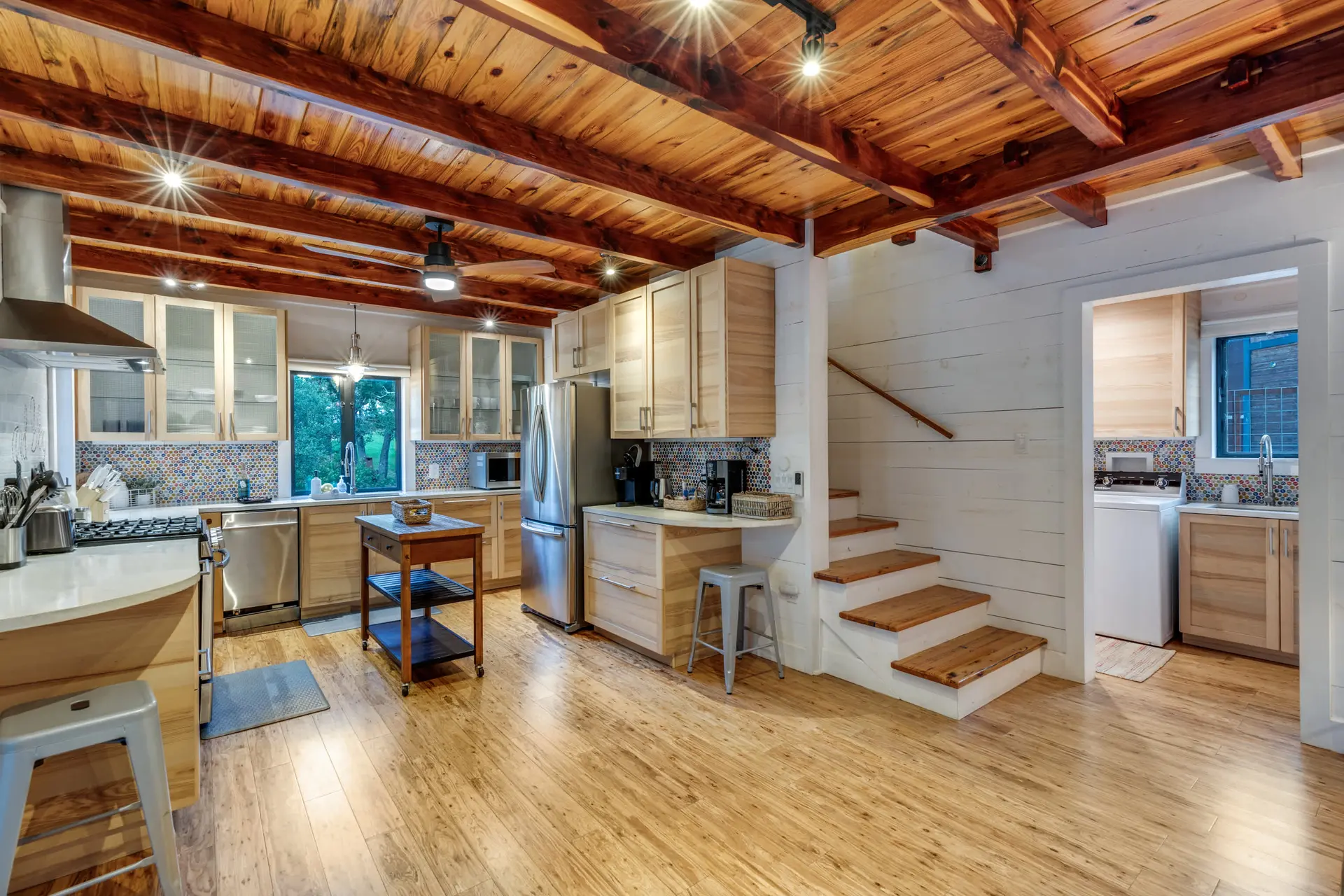 A rustic kitchen with wood-paneled ceiling, stainless steel appliances, and wooden stairs.