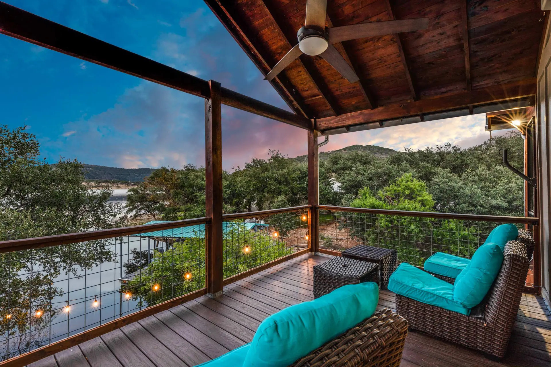 Covered deck with wicker furniture overlooking a lake at sunset.