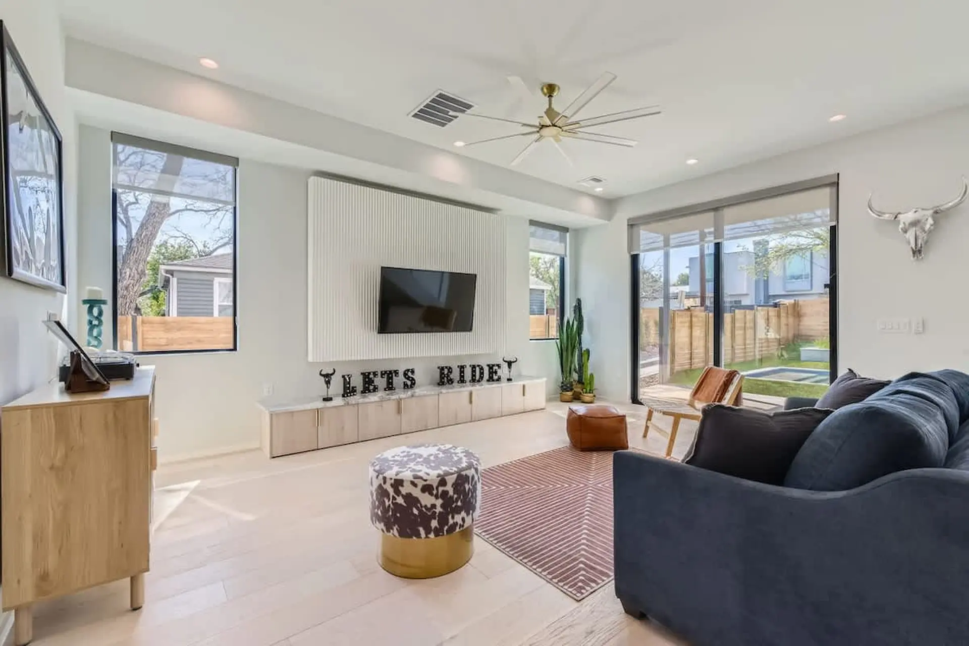 A living room with a TV, sofa, cowhide ottoman, and large windows.