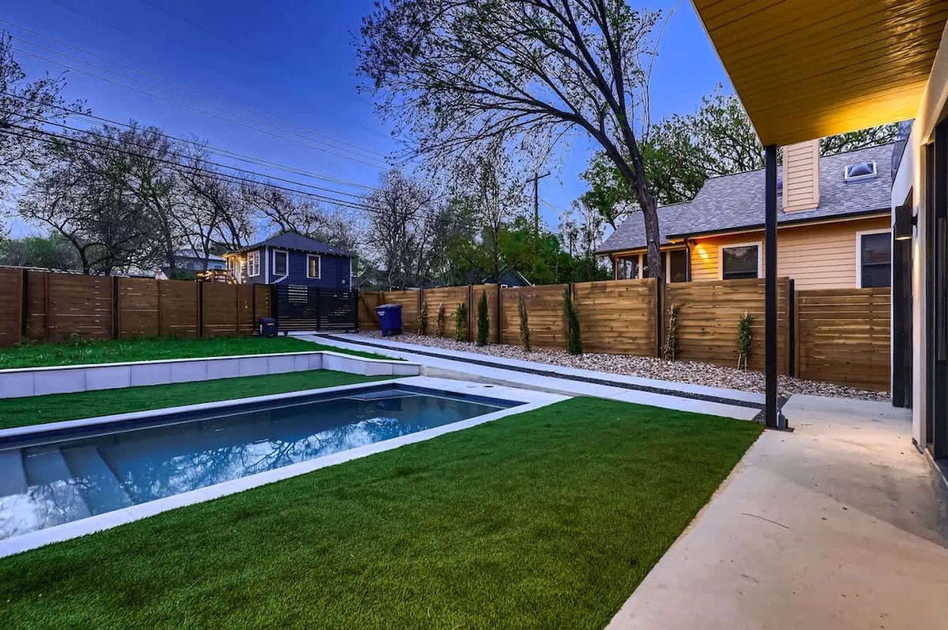 A backyard with a pool, artificial turf, and a wooden fence at dusk.