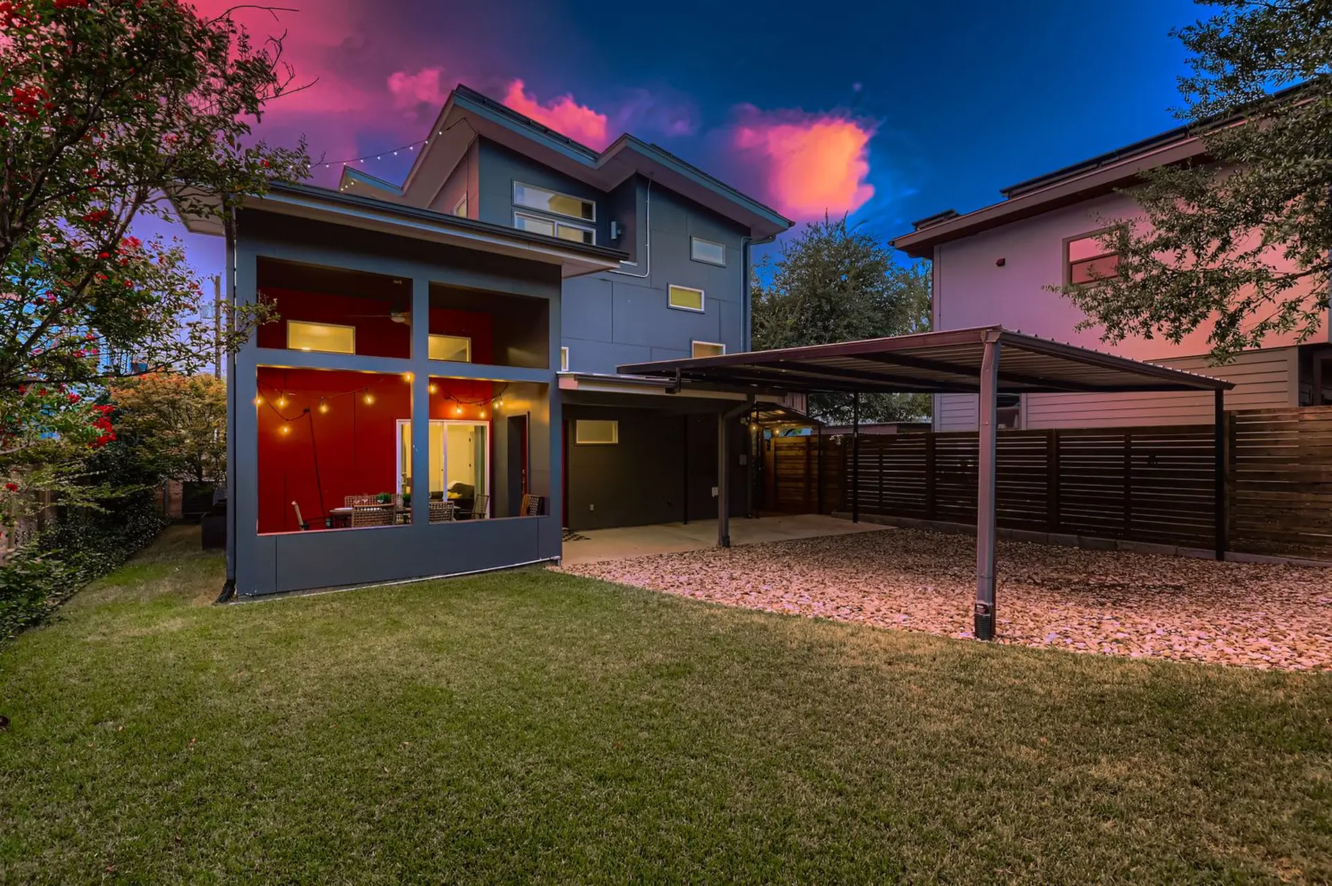 Modern home with covered carport, patio with string lights, and lush green lawn at twilight.