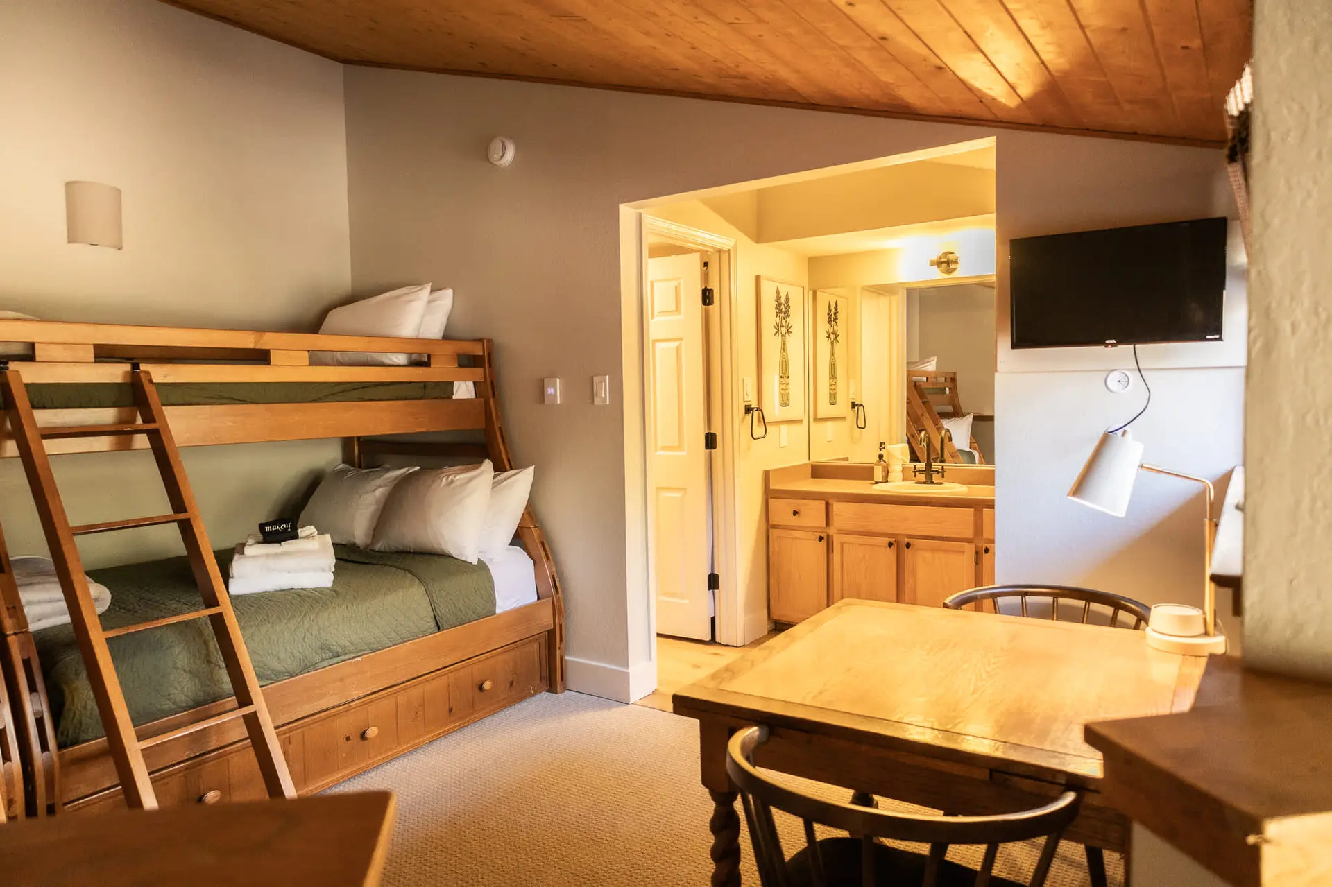 A rustic room features bunk beds, a vanity, a TV, and a wooden table with chairs.