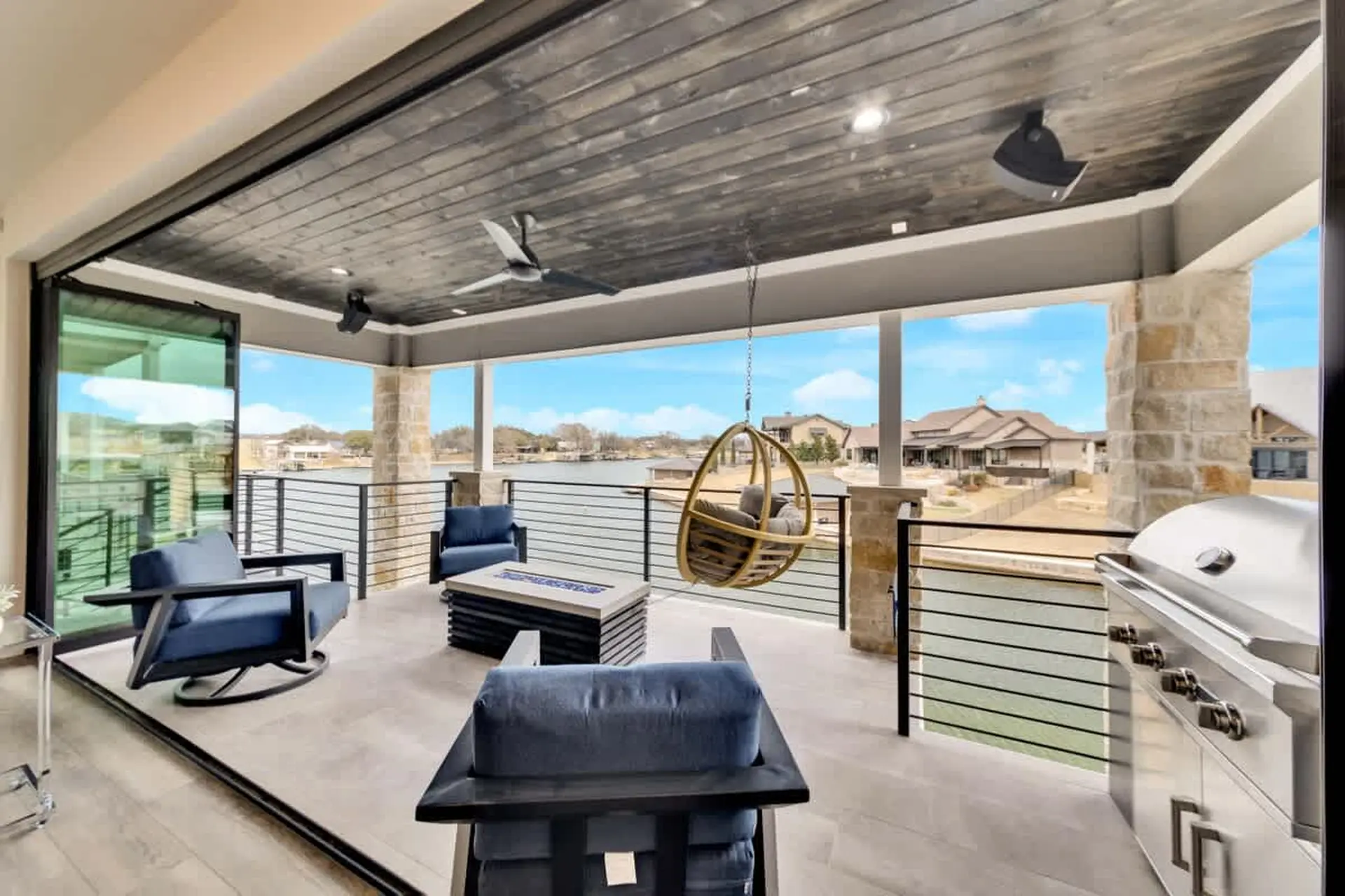 Covered patio with outdoor seating, a fire pit, swing chair, and grill overlooking a lake.