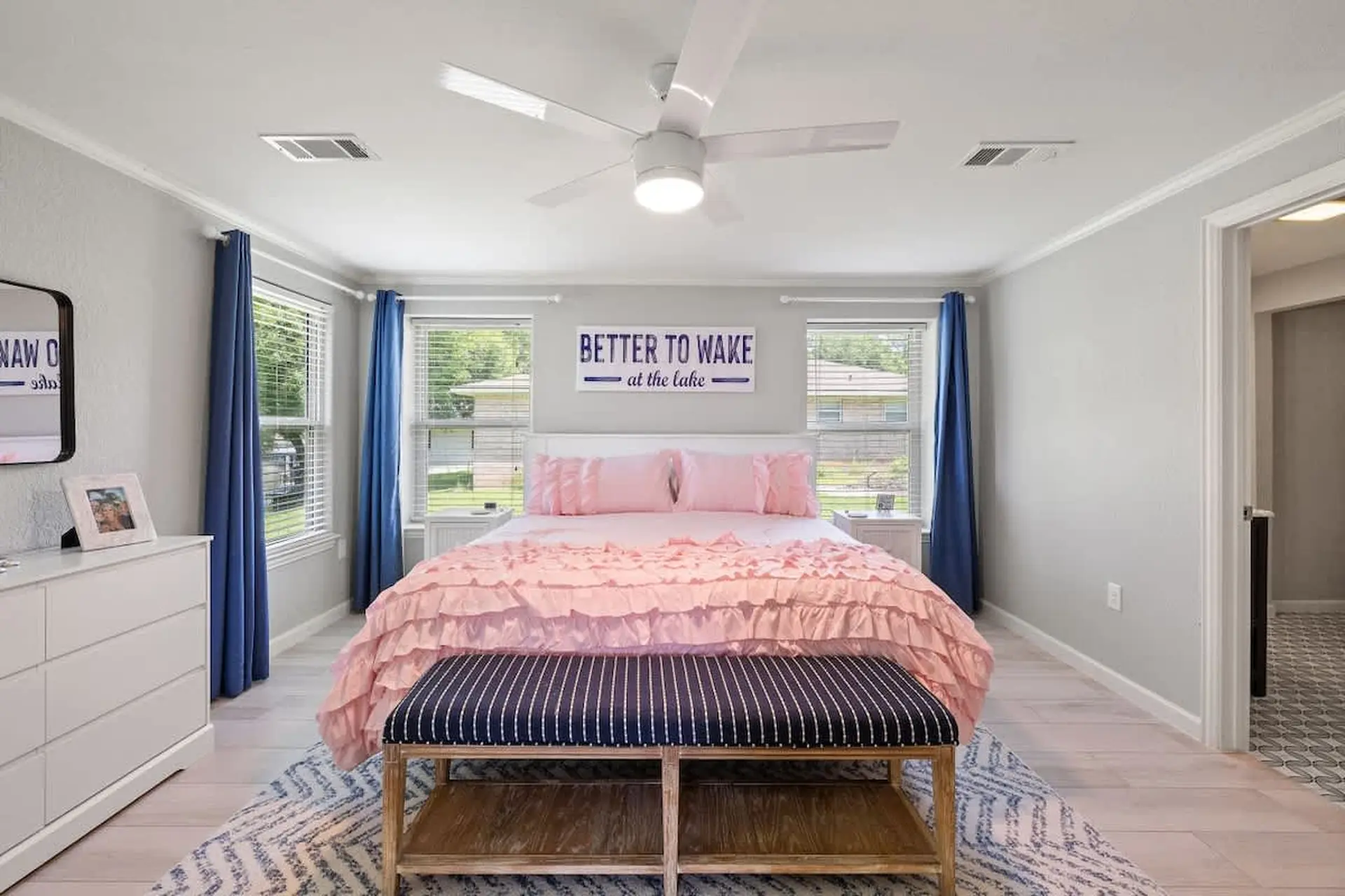 A bedroom with a pink ruffled bedspread, a blue and white striped bench, and blue curtains.