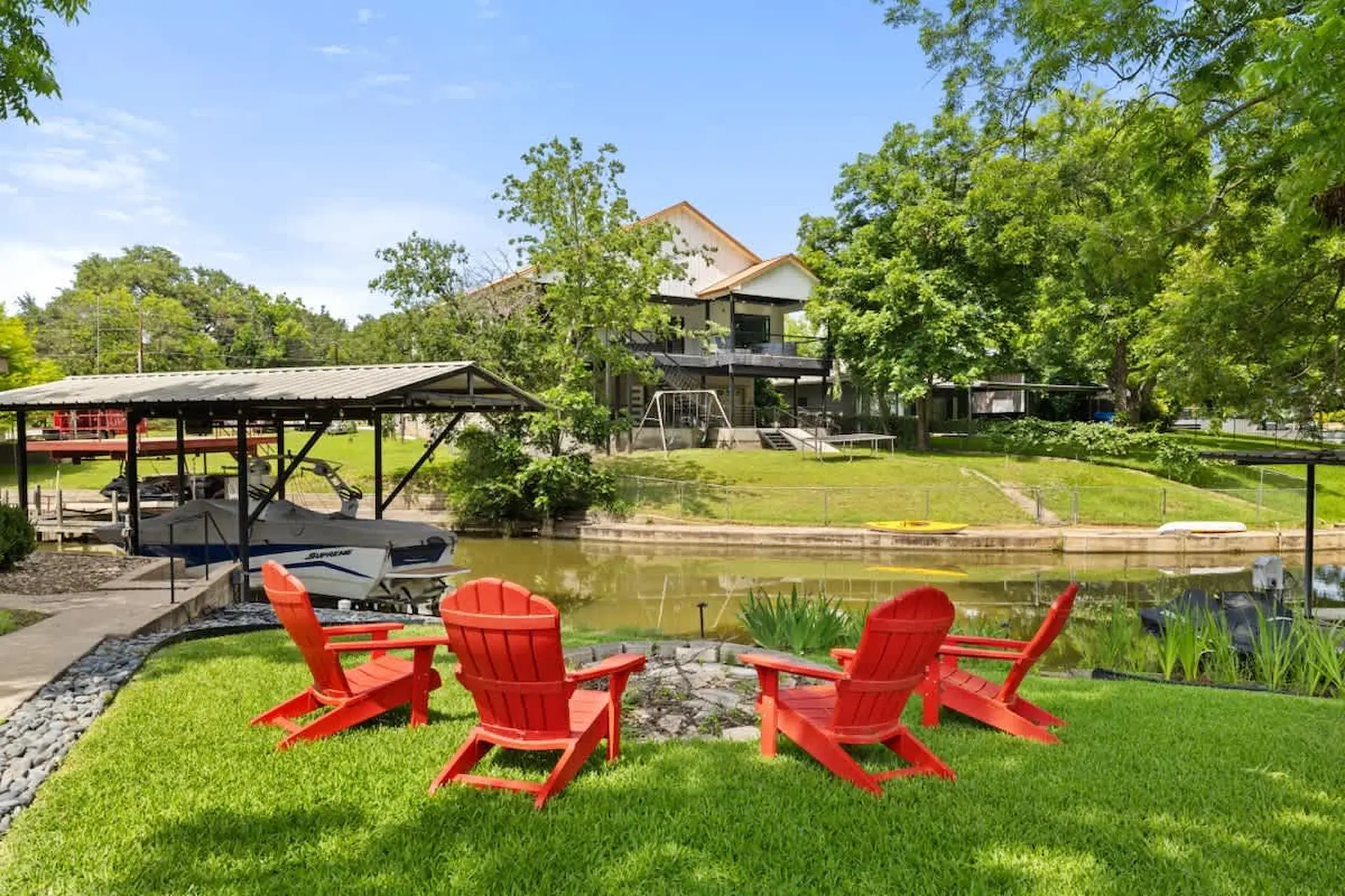 Red Adirondack chairs on a grassy lawn beside a canal with a boat dock and house.