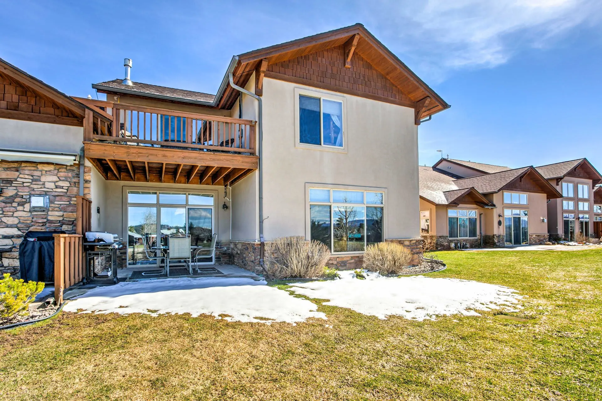 Back view of a beige house with a wooden deck and stone accents, featuring a grill and outdoor seati