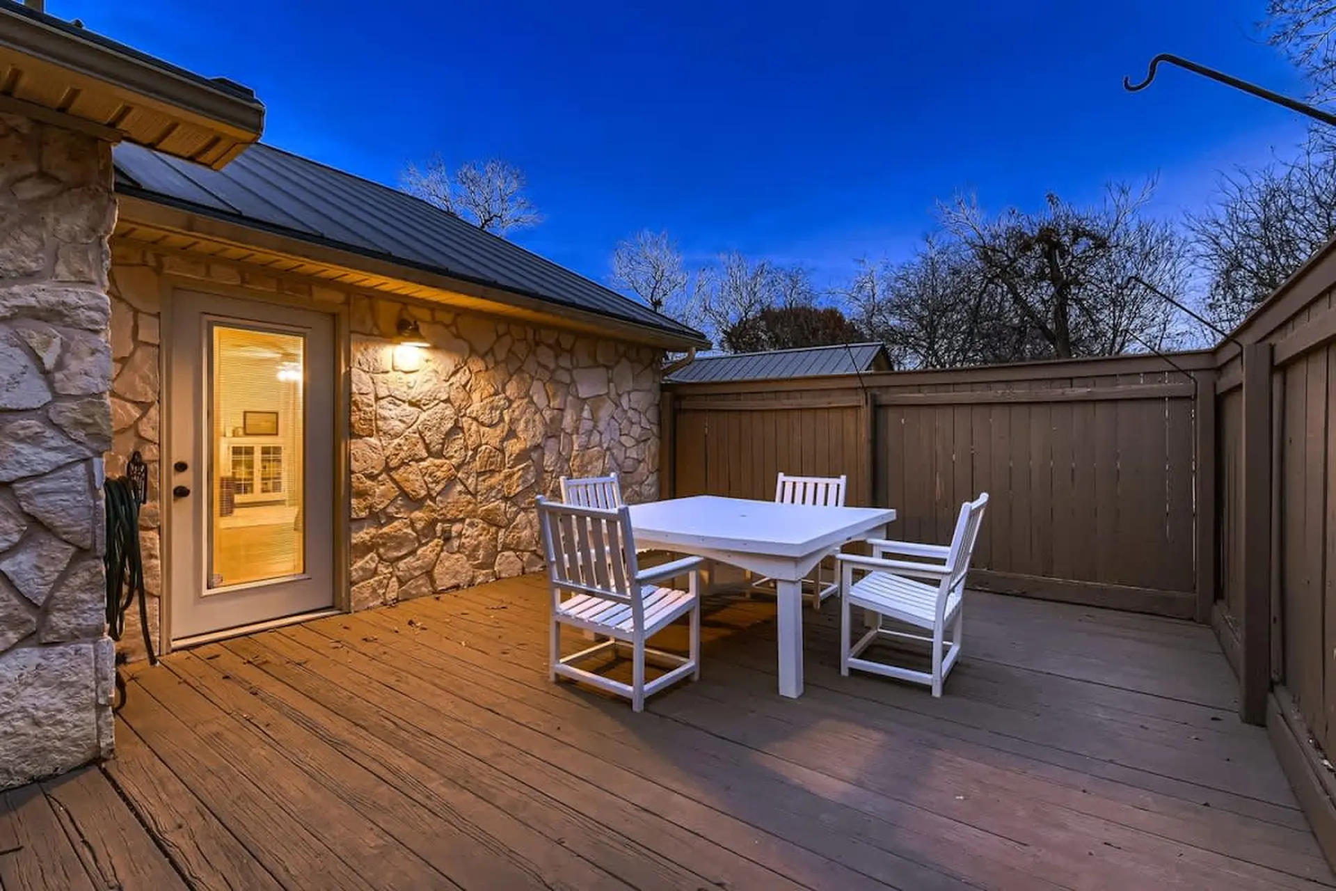 Outdoor patio with white dining set against stone wall and wooden fence under a twilight sky.