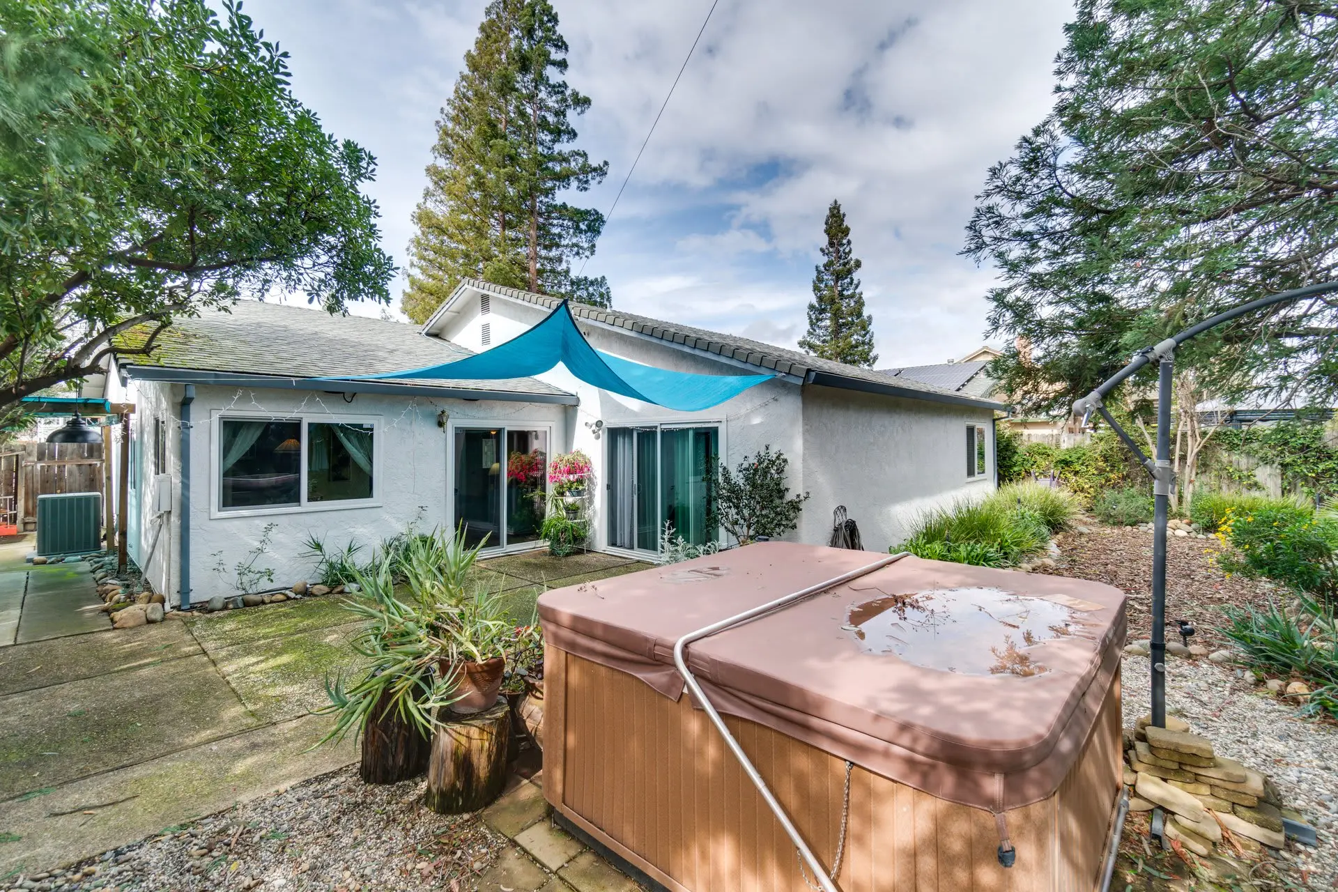 A backyard with a hot tub, concrete patio, gravel, plants, and a white stucco house.