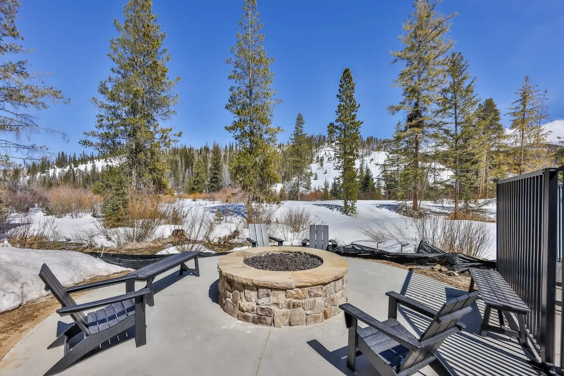 Outdoor patio with a stone fire pit and chairs, surrounded by pine trees and snow-covered hills.