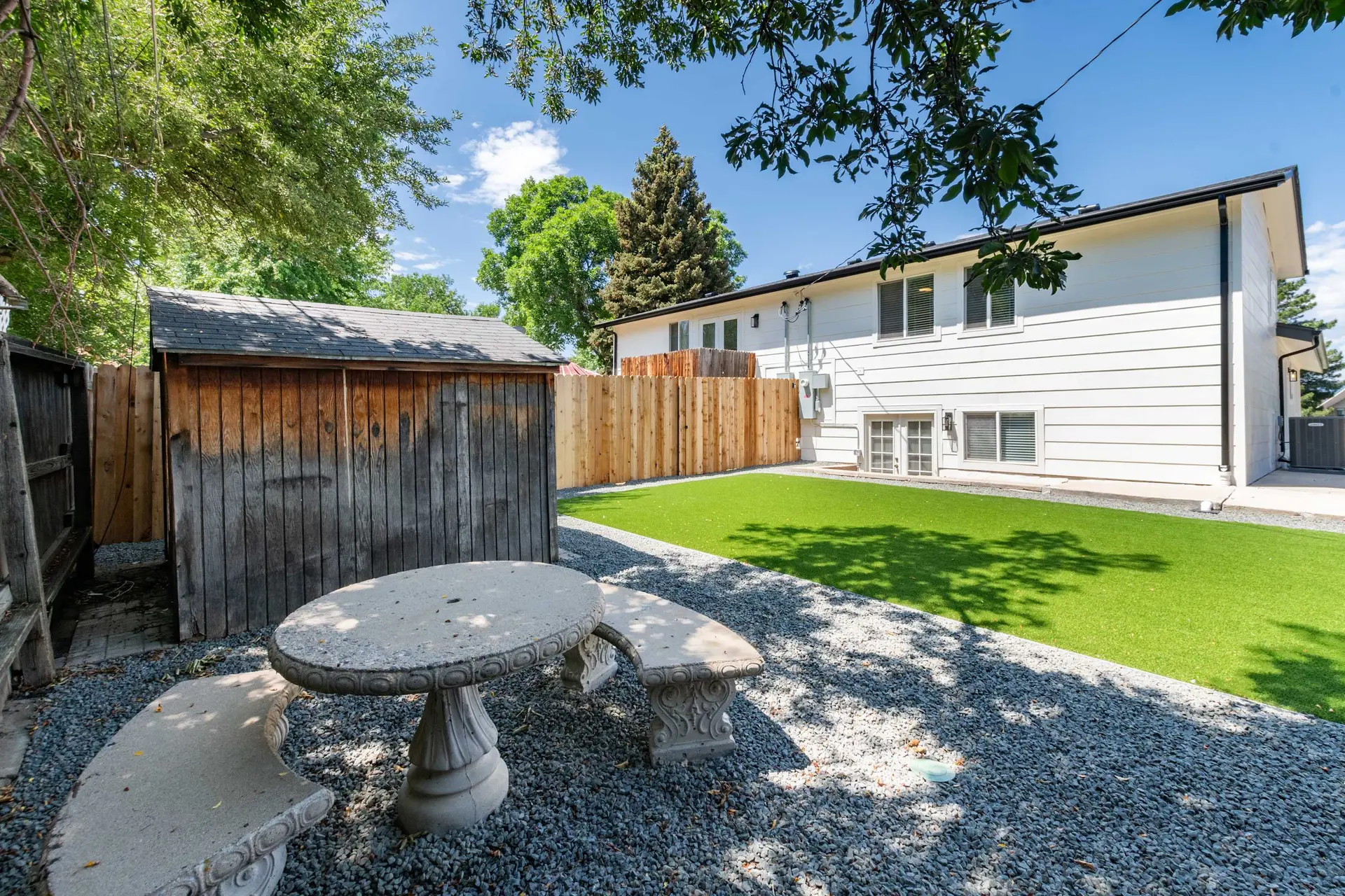 Backyard with white house, green turf, wooden shed, and stone table with benches.