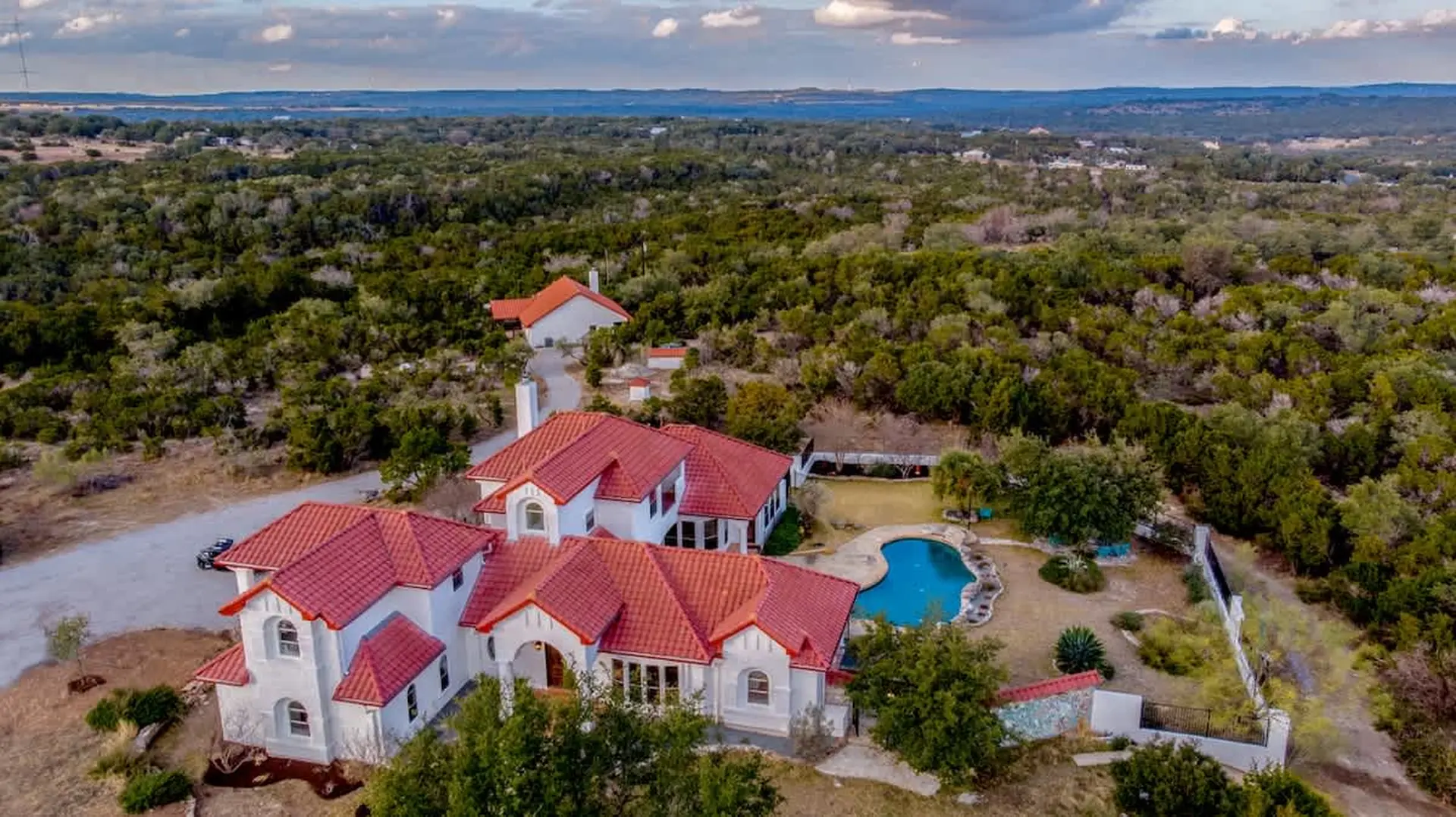 Aerial view of a large white house with red tile roof and pool in a wooded area.