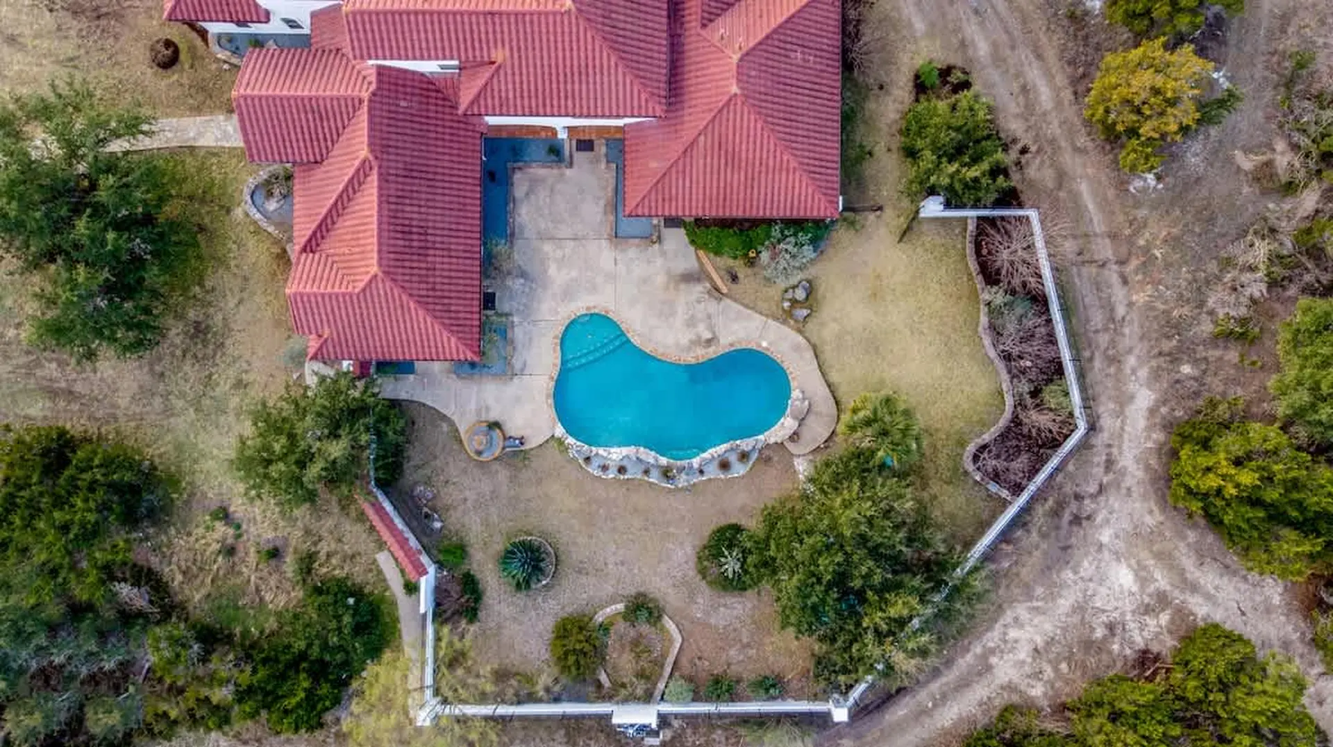 An aerial view of a red-roofed house with a kidney-shaped pool and surrounding greenery.