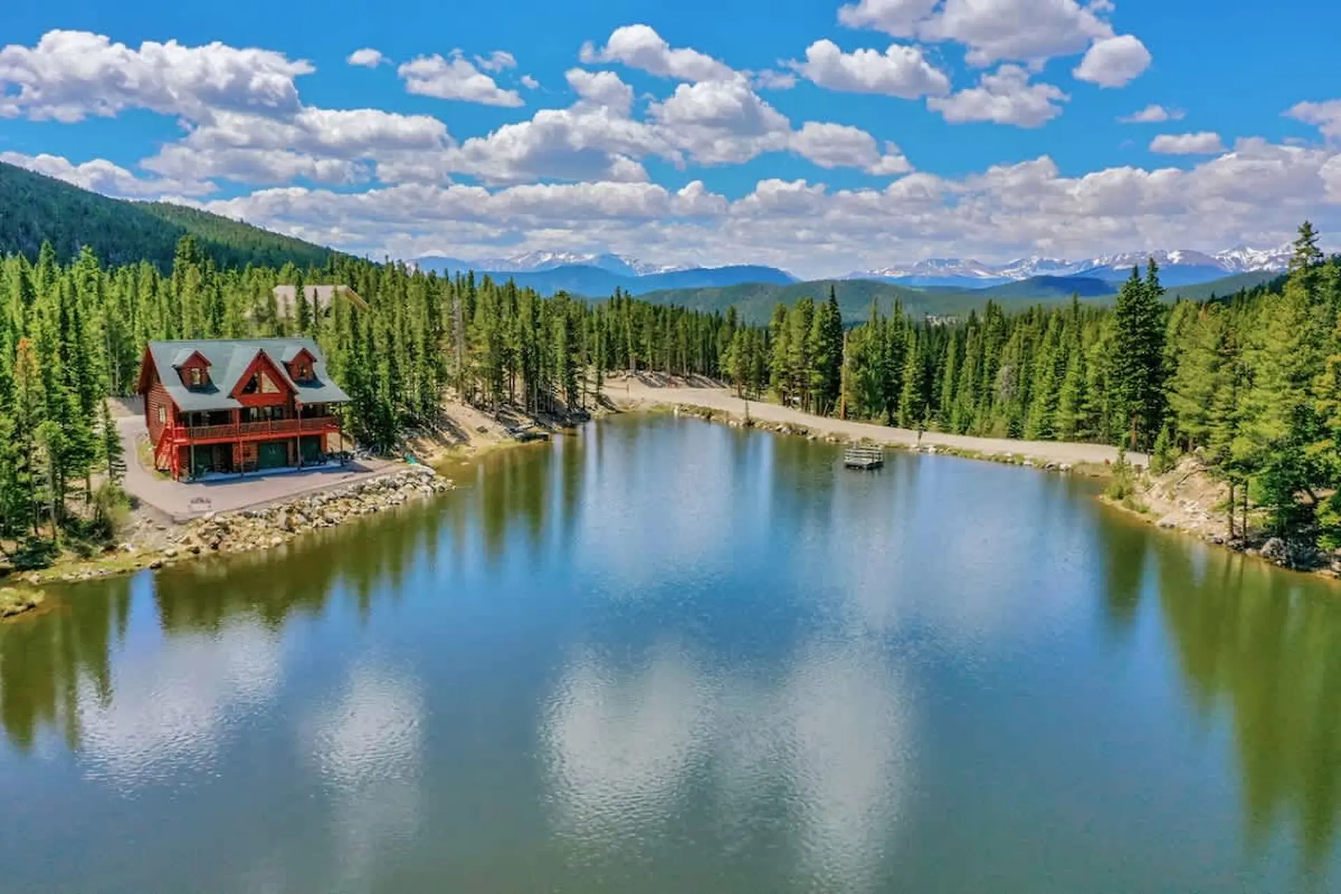 A log cabin sits by a lake, surrounded by pine trees and mountains.