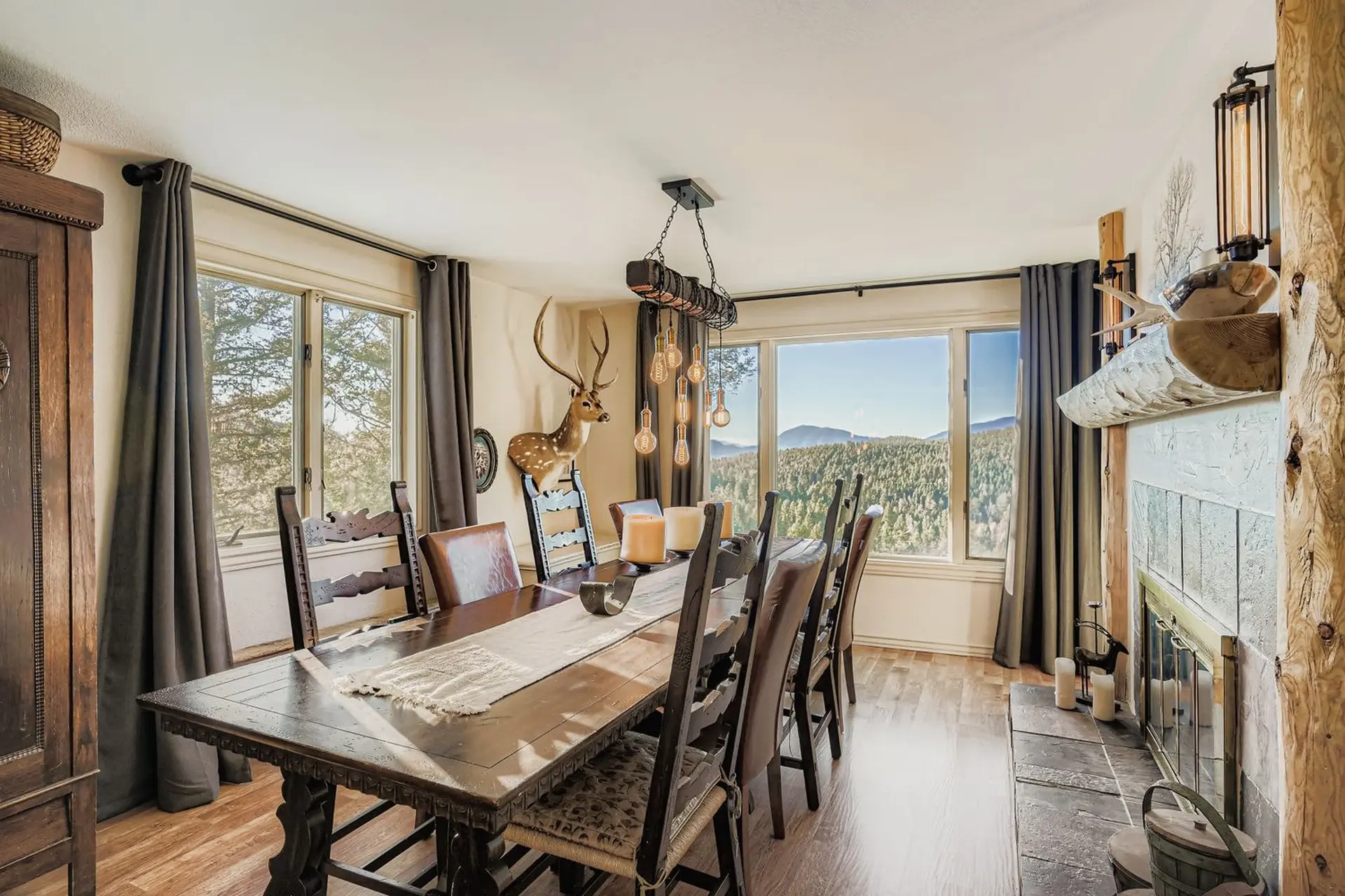 A rustic dining room with a large wooden table, deer mount, and fireplace overlooking a forest.