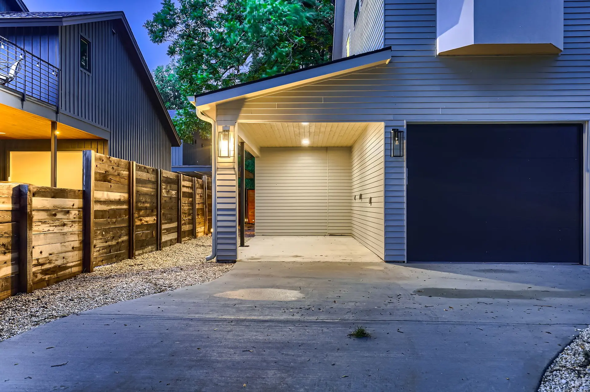 An exterior shot of a modern home featuring a covered carport, a dark garage door, and a wooden fenc