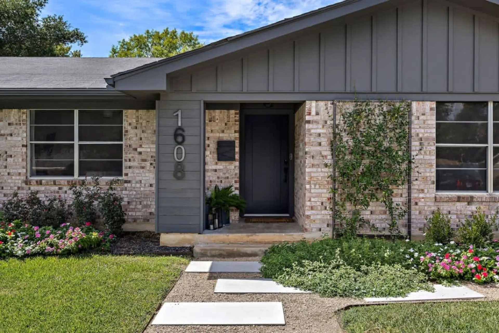 Front exterior of a brick house with a dark door, large windows, and lush landscaping.