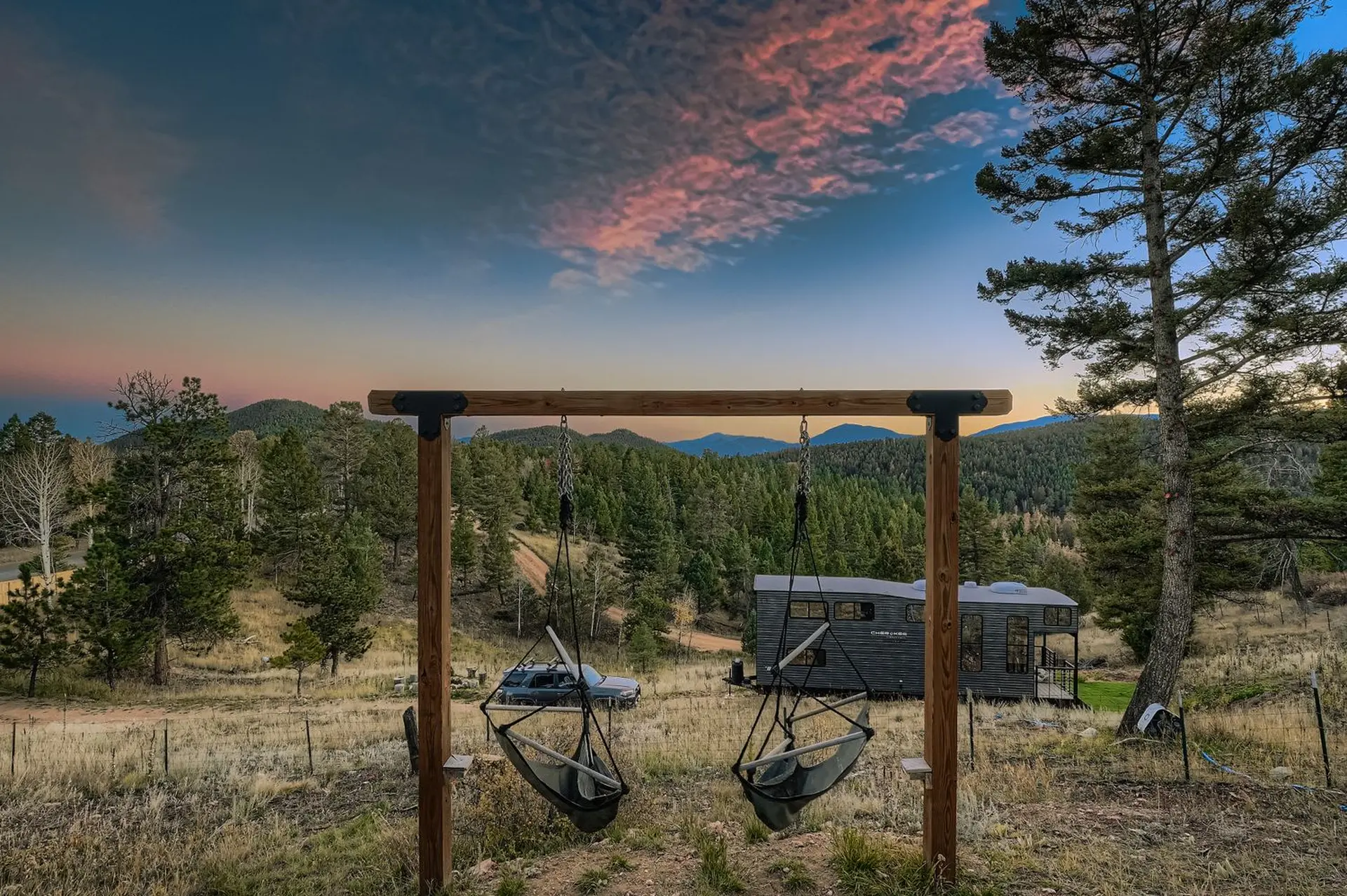 Two swings hang in front of a tiny home and wooded landscape at sunset.
