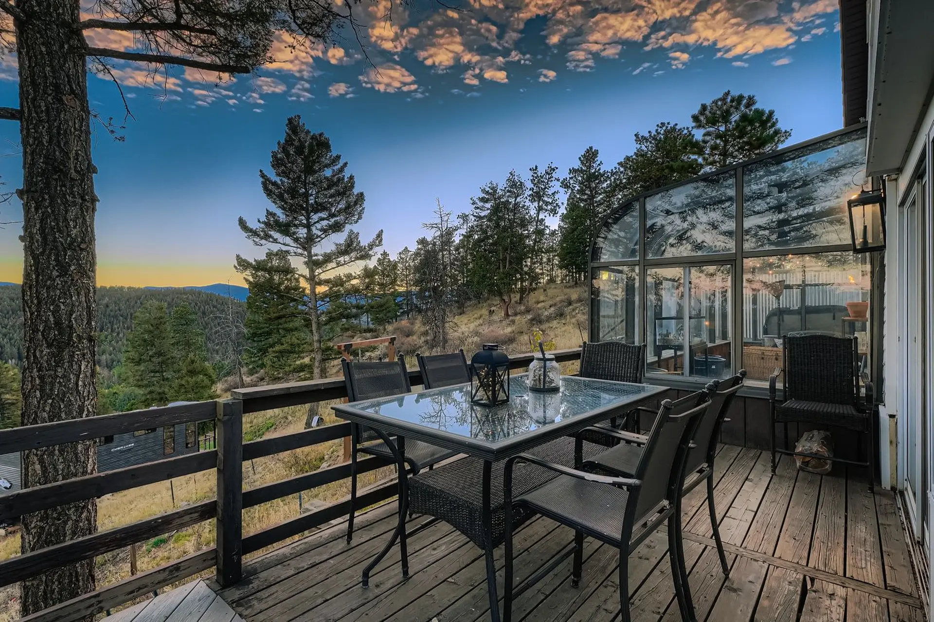 Deck with table and chairs overlooks forest and greenhouse at sunset.