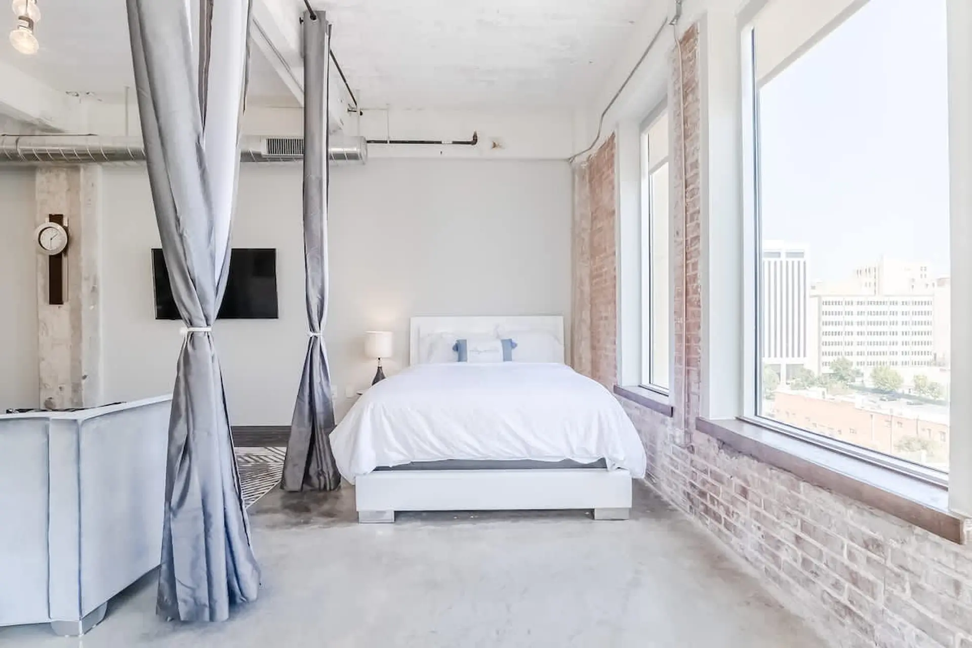 A loft bedroom with a white bed, exposed brick walls, and large windows overlooking a city.
