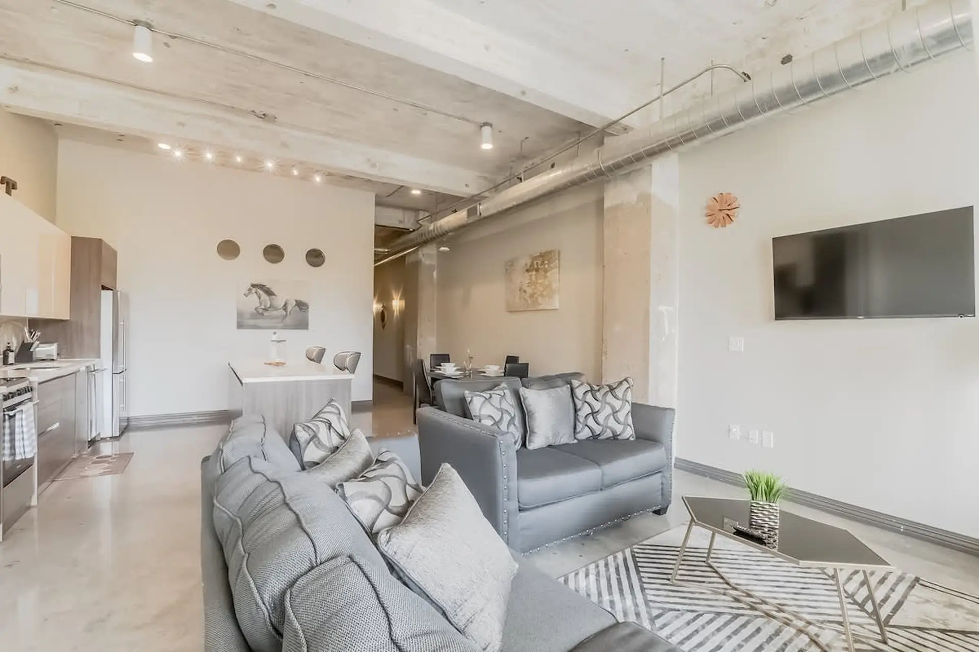 Modern loft living room with open-plan kitchen, gray sofas, and a television mounted on the wall.