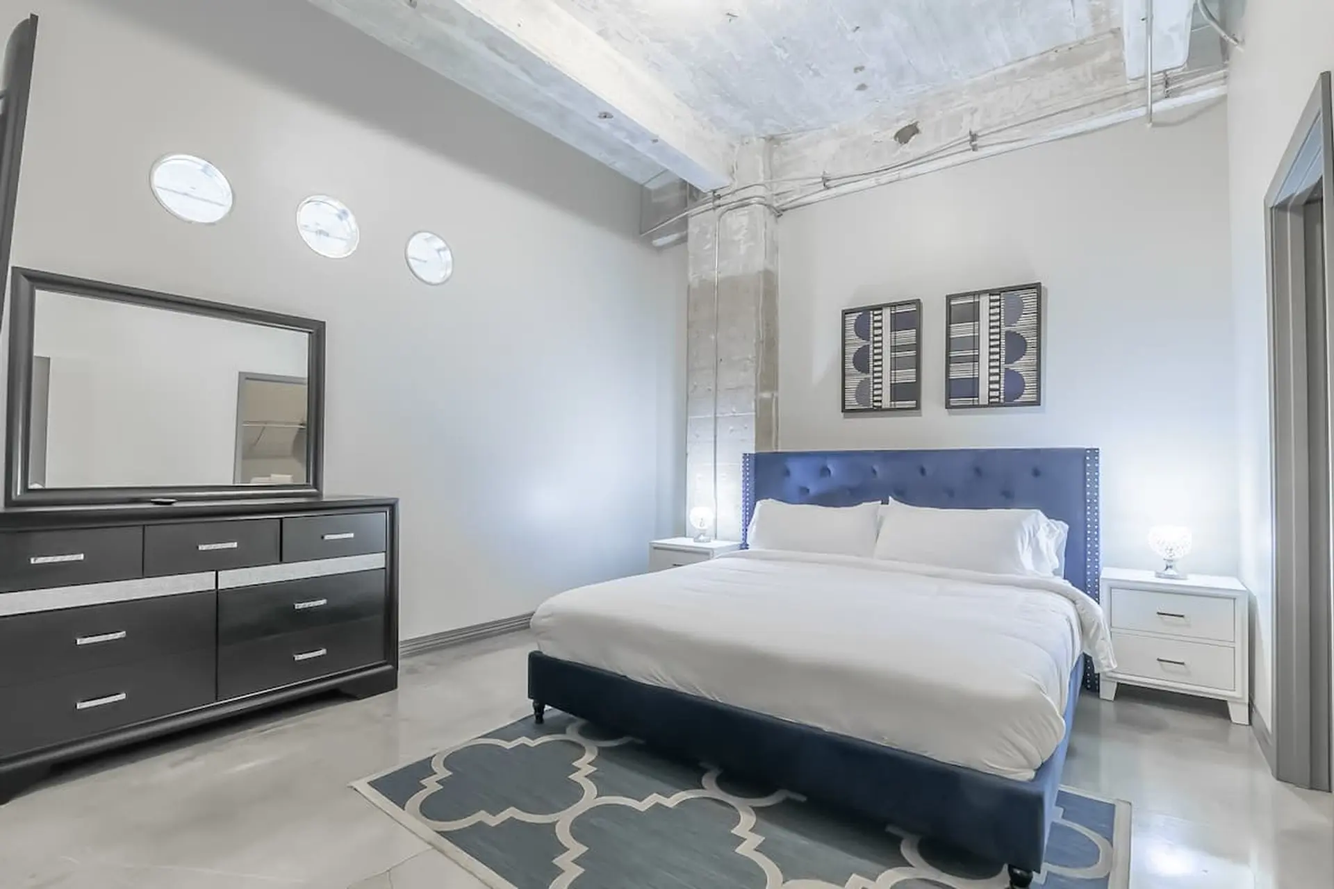 A modern bedroom with a blue tufted bed, dresser, and abstract art.