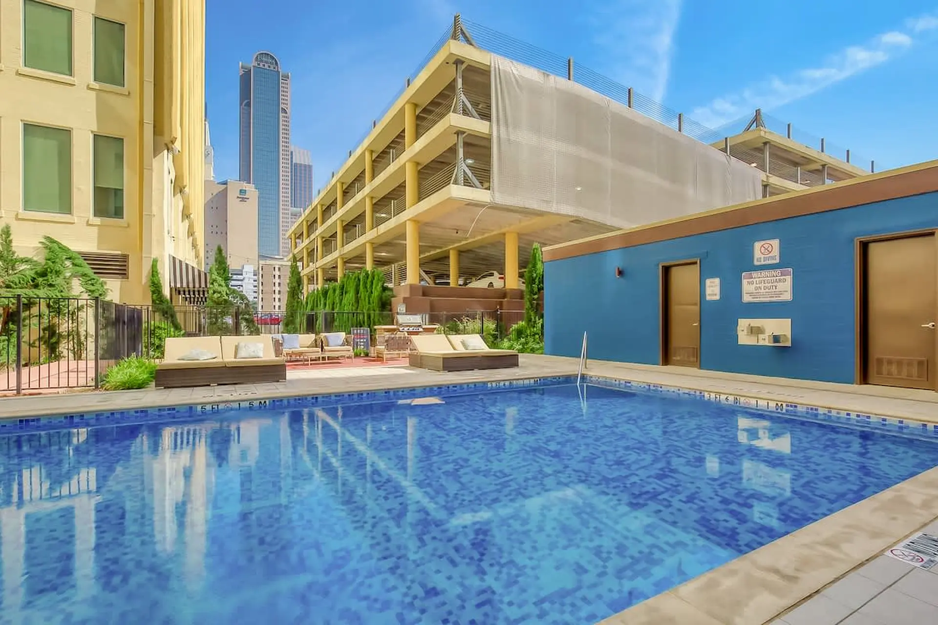 A blue tiled swimming pool with lounge chairs in the background, a parking garage, and a tall skyscr