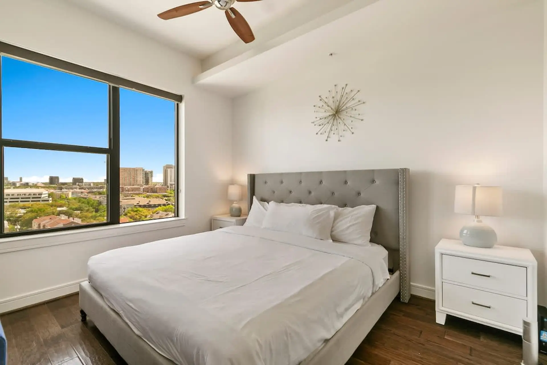 A bedroom with a large window overlooking a city, a king-size bed, and a nightstand.
