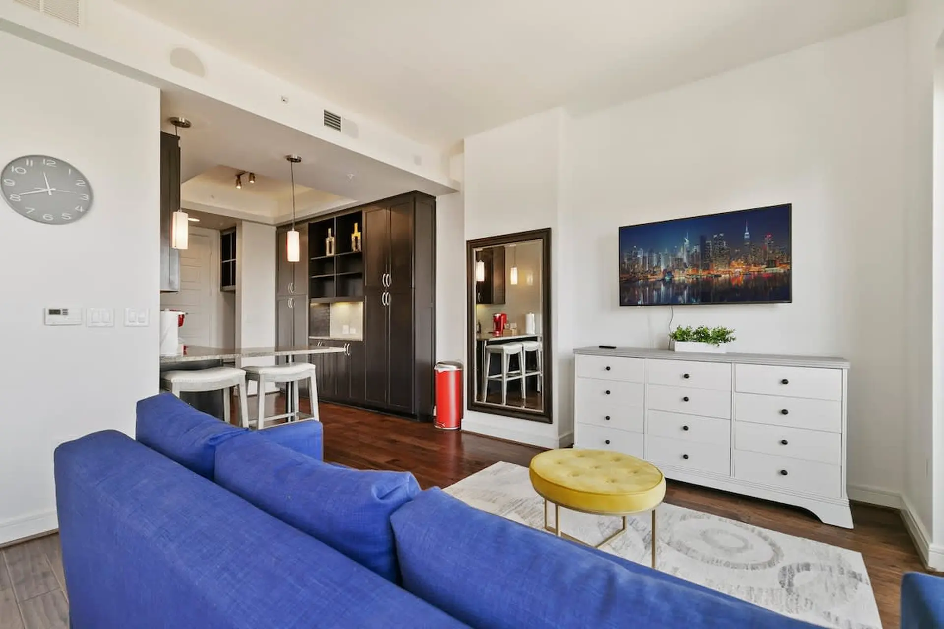 A blue couch faces a dresser with a TV and a yellow ottoman in a modern living room.