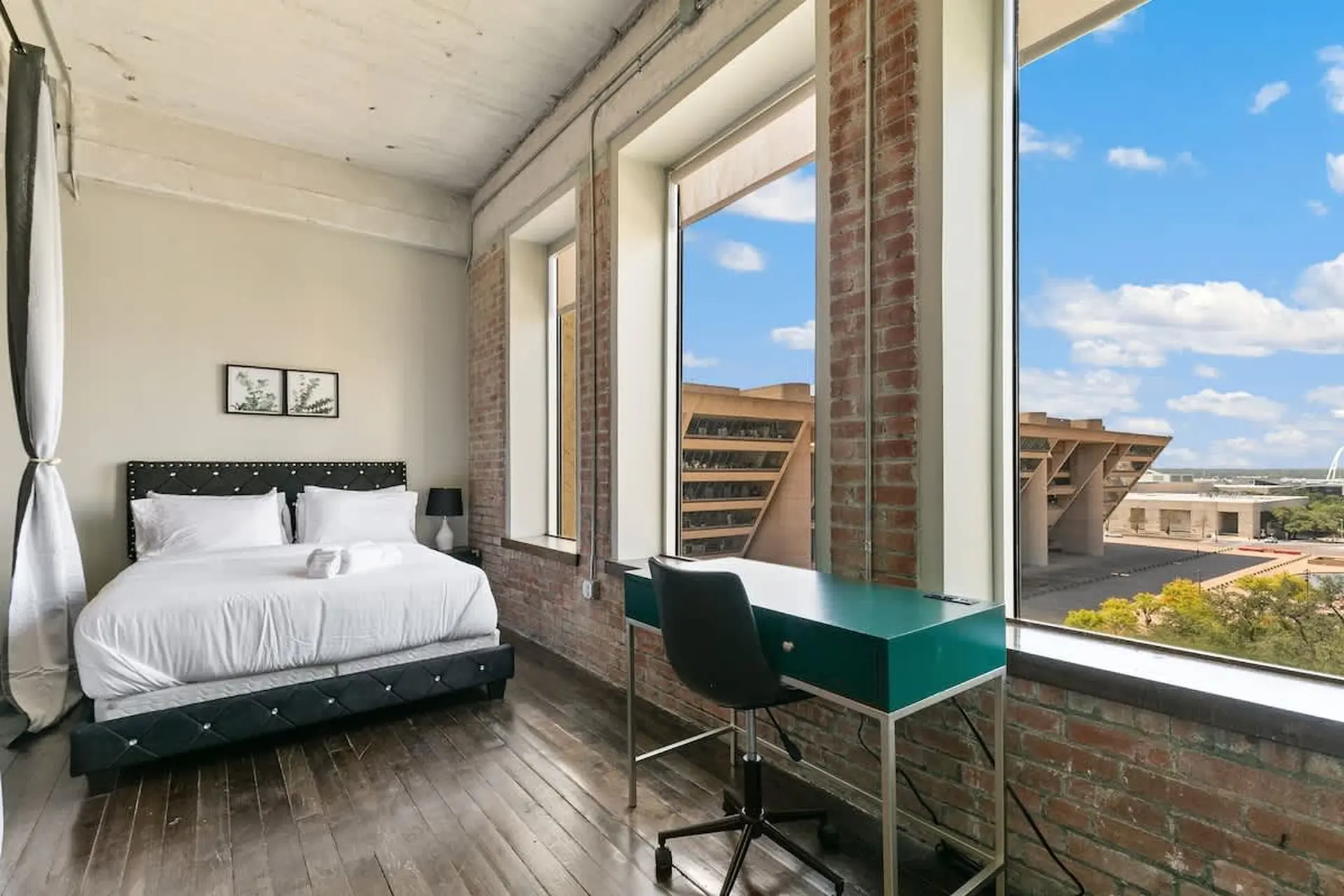 Bedroom with a bed, desk and large windows overlooking a city skyline.
