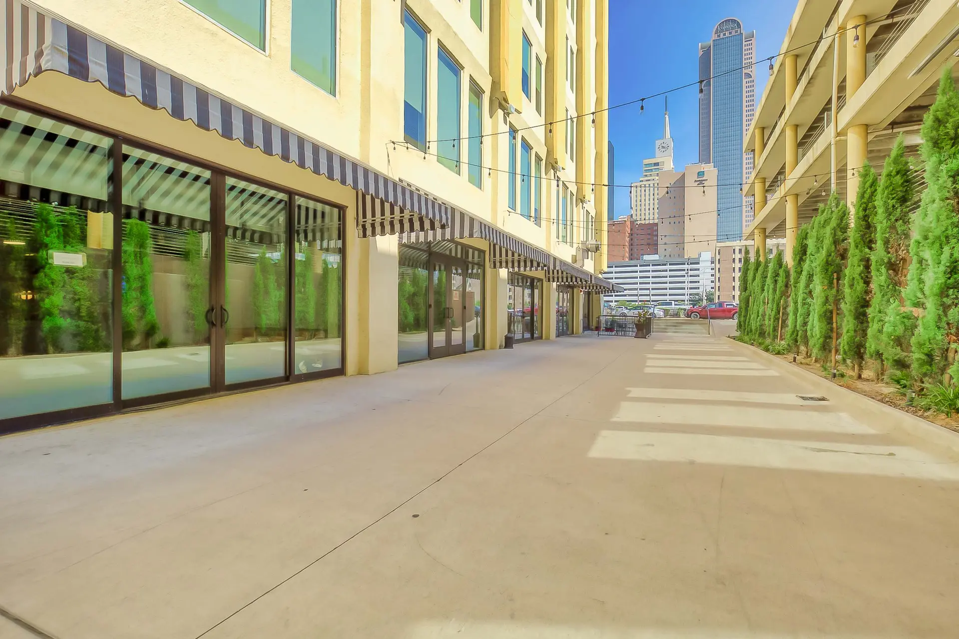 A sunlit pedestrian walkway lined with trees and buildings, with a cityscape in the background.