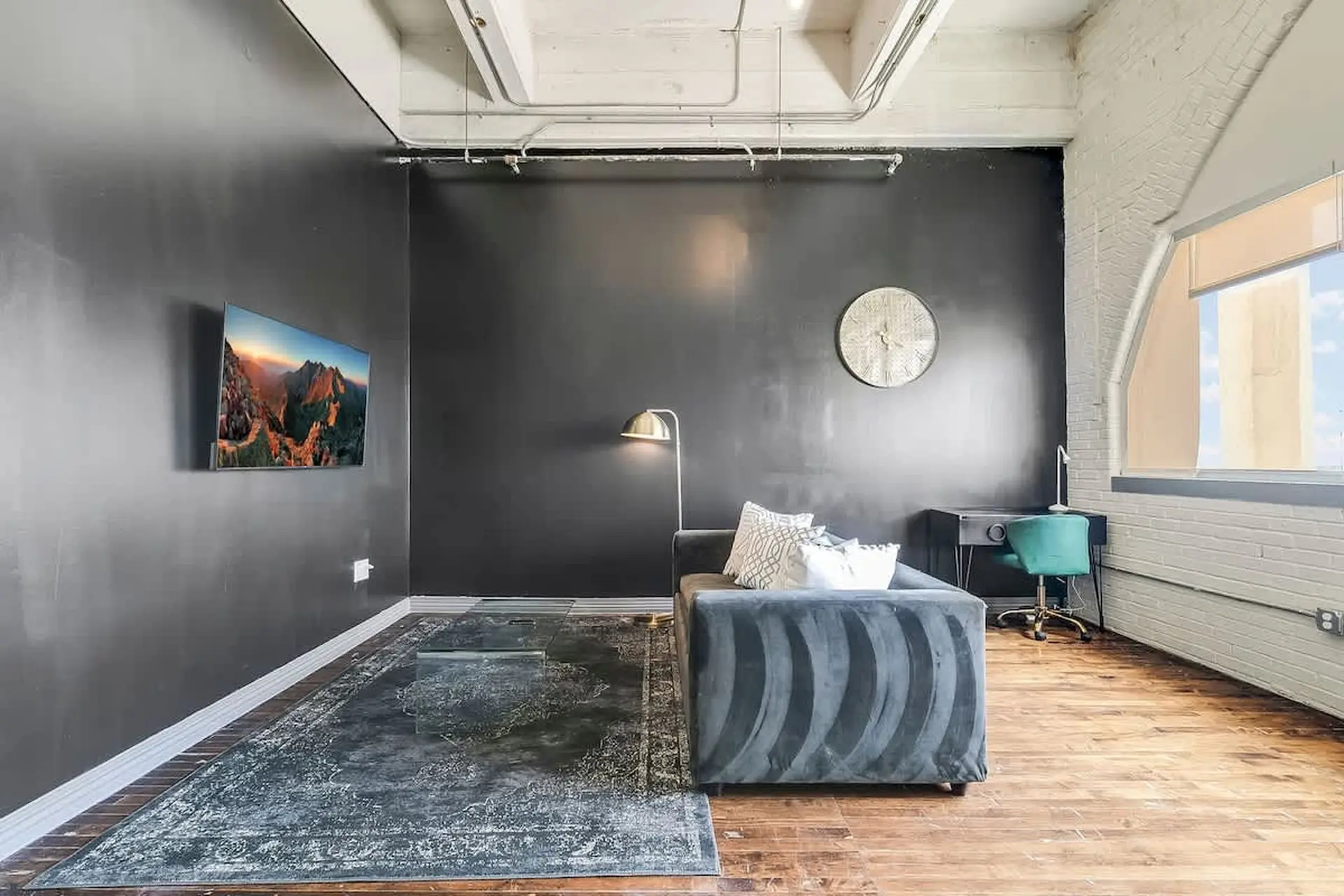 Modern loft living room with a gray sofa, glass coffee table, and mountain artwork on the wall.