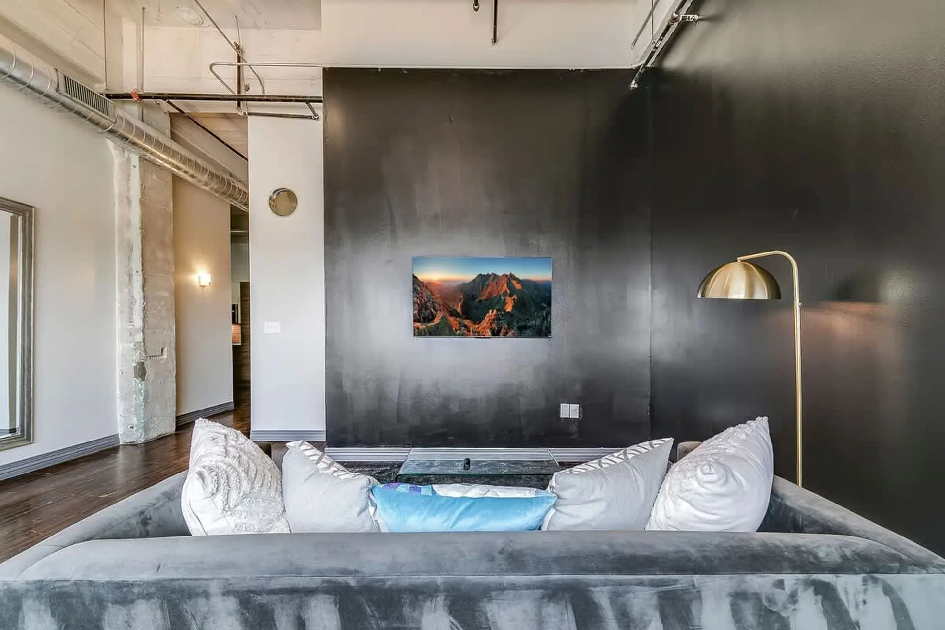 A plush gray sofa faces a dark wall with a mountain landscape painting and a golden floor lamp.