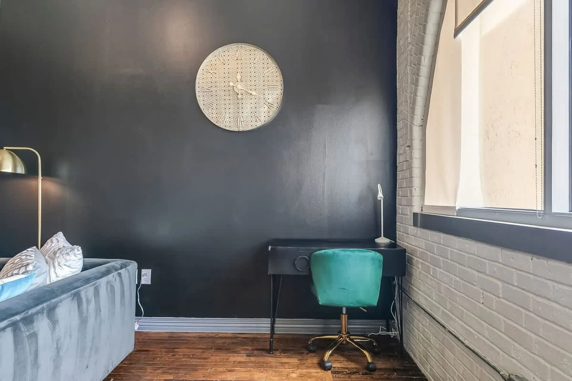 A black desk with a teal chair sits against a black wall with a clock and a gold floor lamp.