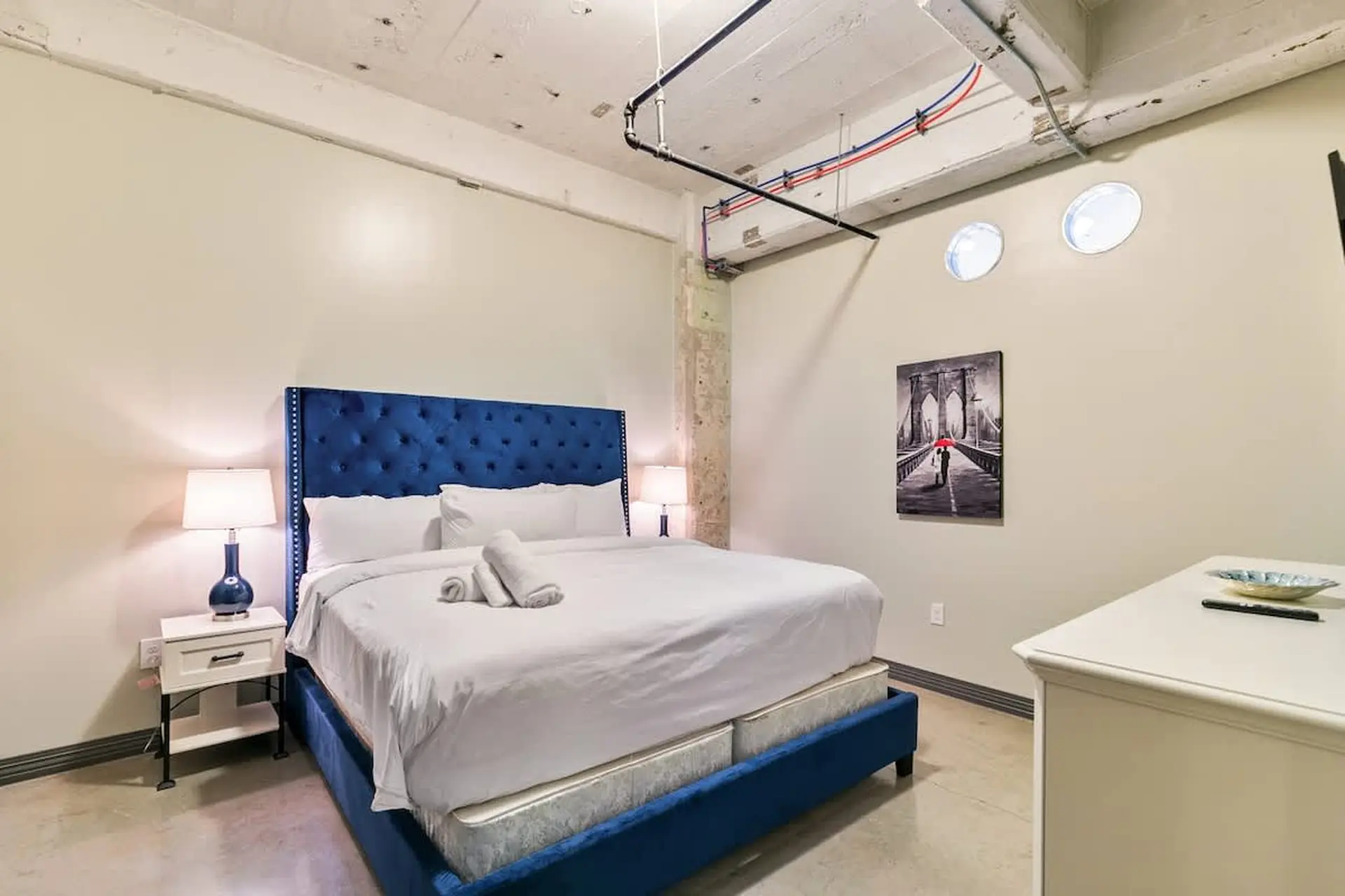 A bedroom with a blue tufted headboard, white bedding, and industrial-style ceiling pipes.